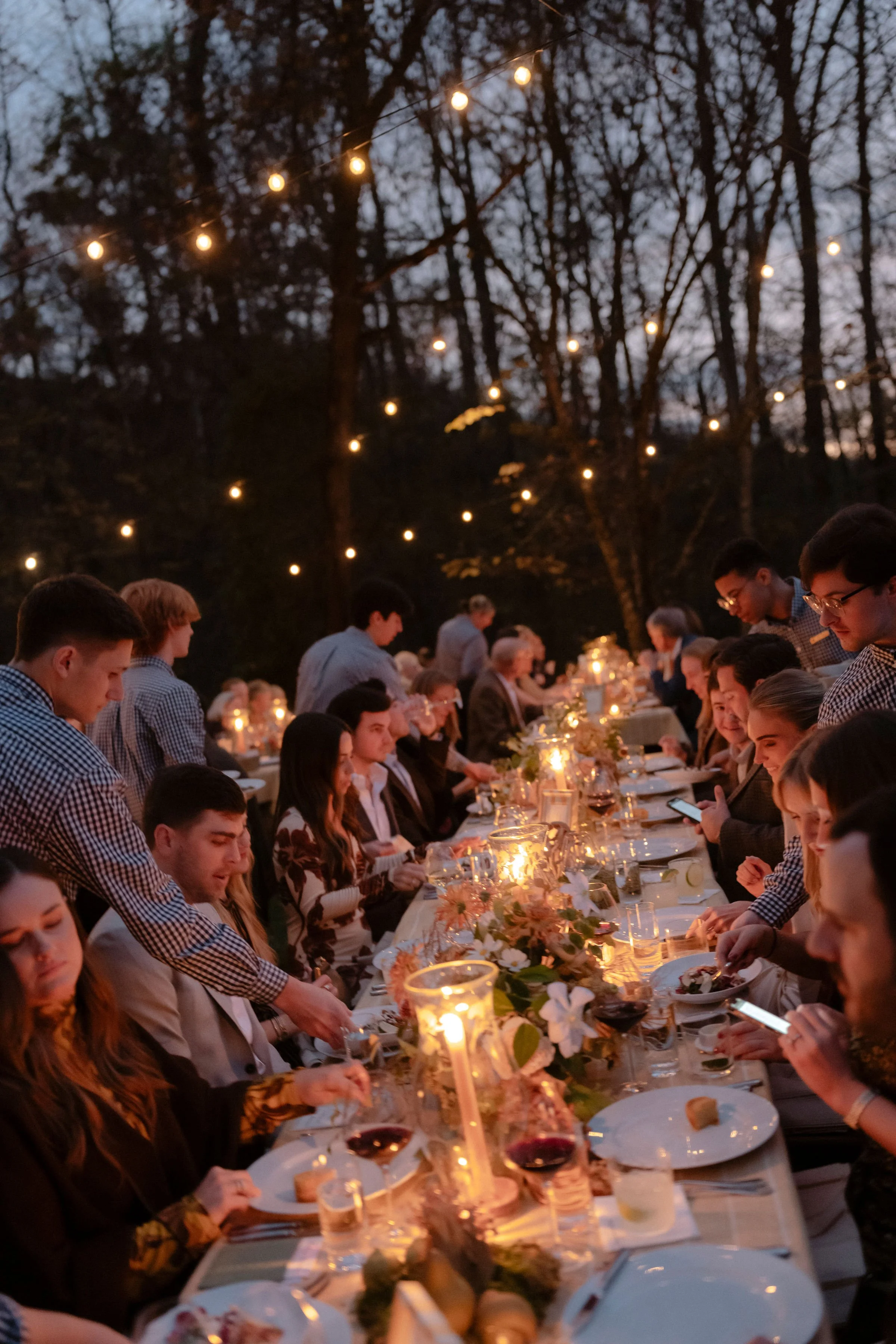Dinner party outdoors at dusk with string lights overhead, long table decorated with flowers, hurricane glass covering candles.