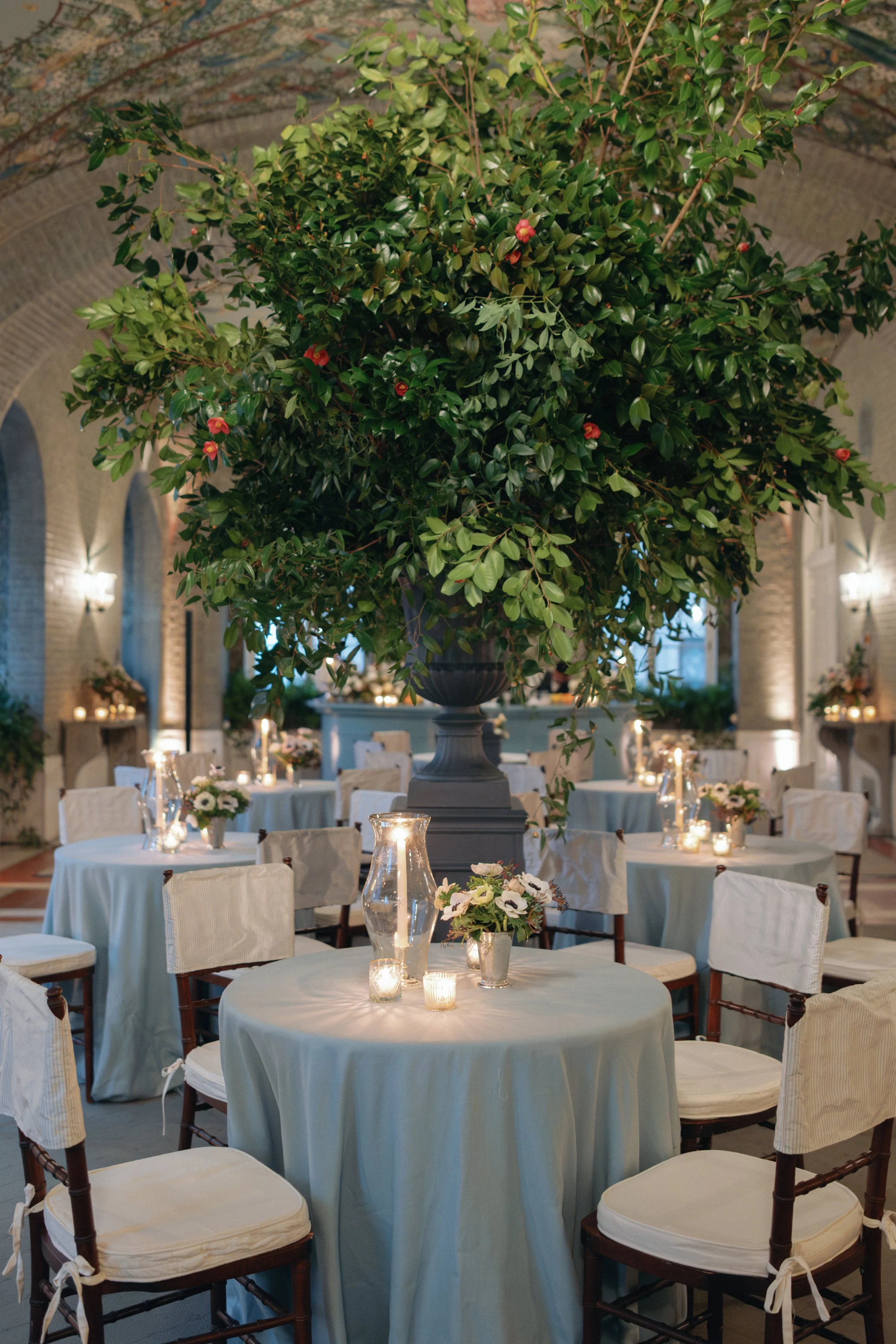Elegant indoor event space with round tables covered in white tablecloths, decorated with candles, vases of flowers, and a large potted plant centerpiece, set in a room with arched brick walls and soft lighting.