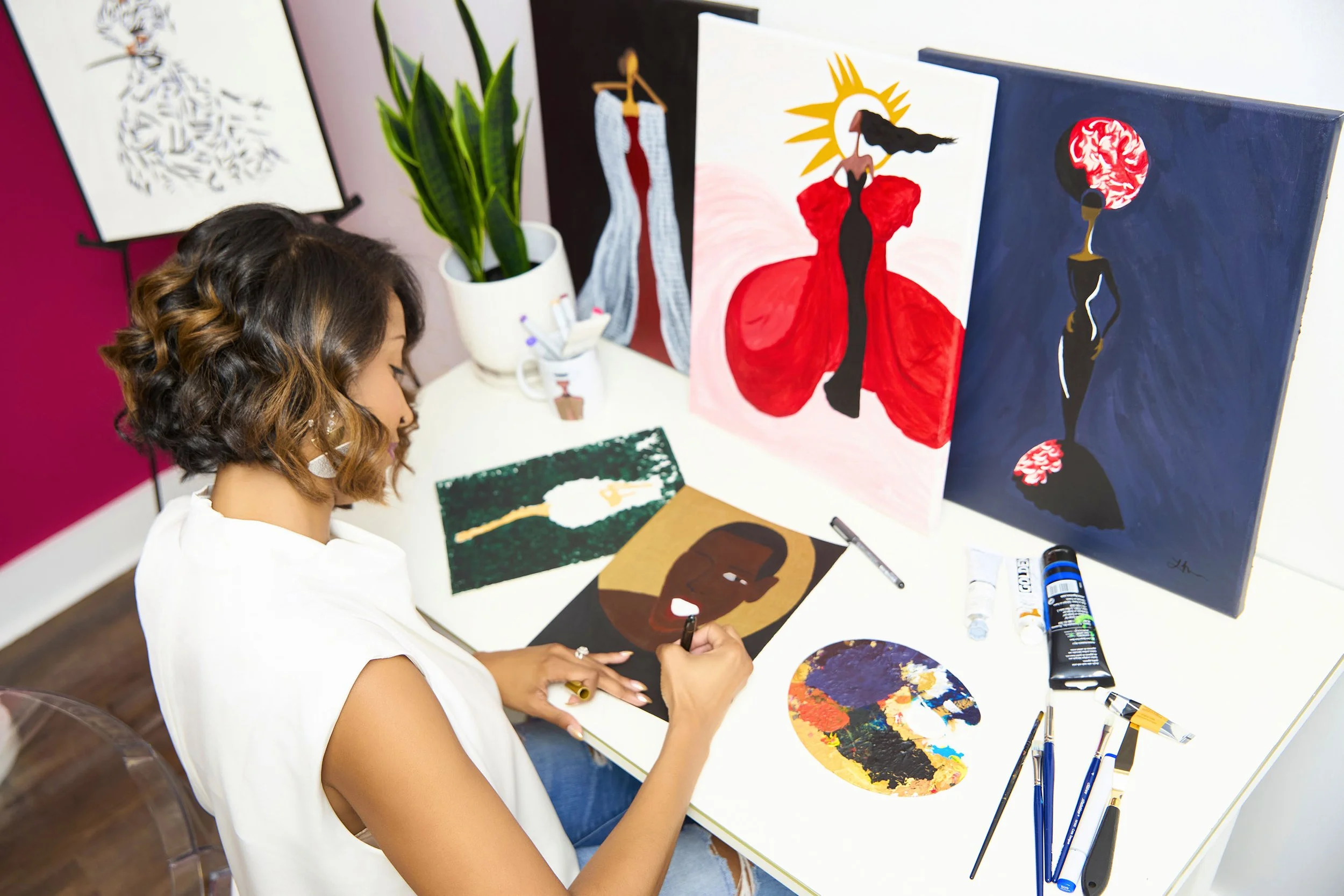 Artist Joelle Monique painting at a table with various artworks displayed, including abstract and fashion-themed designs. Brushes, paints, and a color palette are visible.