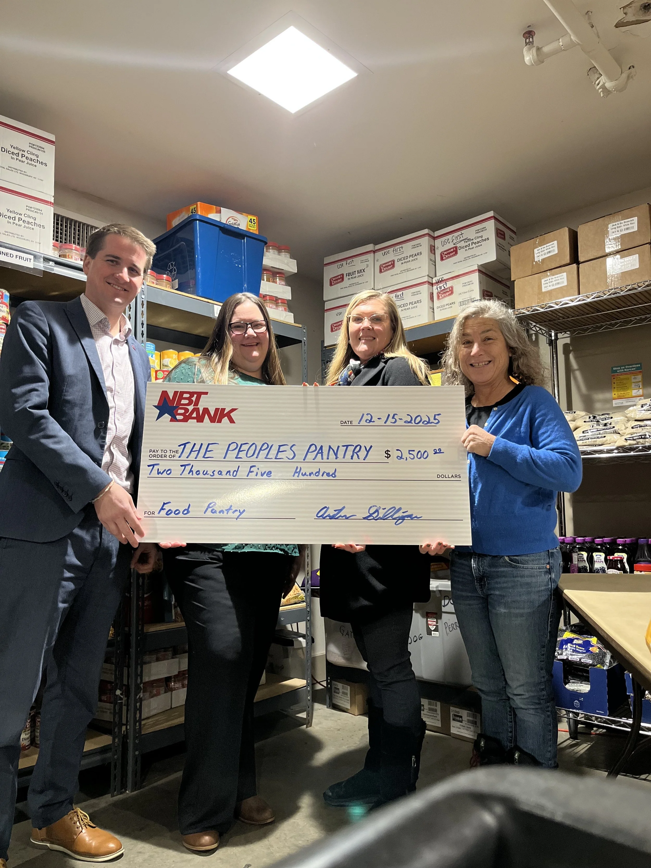 Three people in formal attire holding a large donation check for $2,500 made out to The People's Pantry, standing in front of shelves filled with canned food and other supplies.