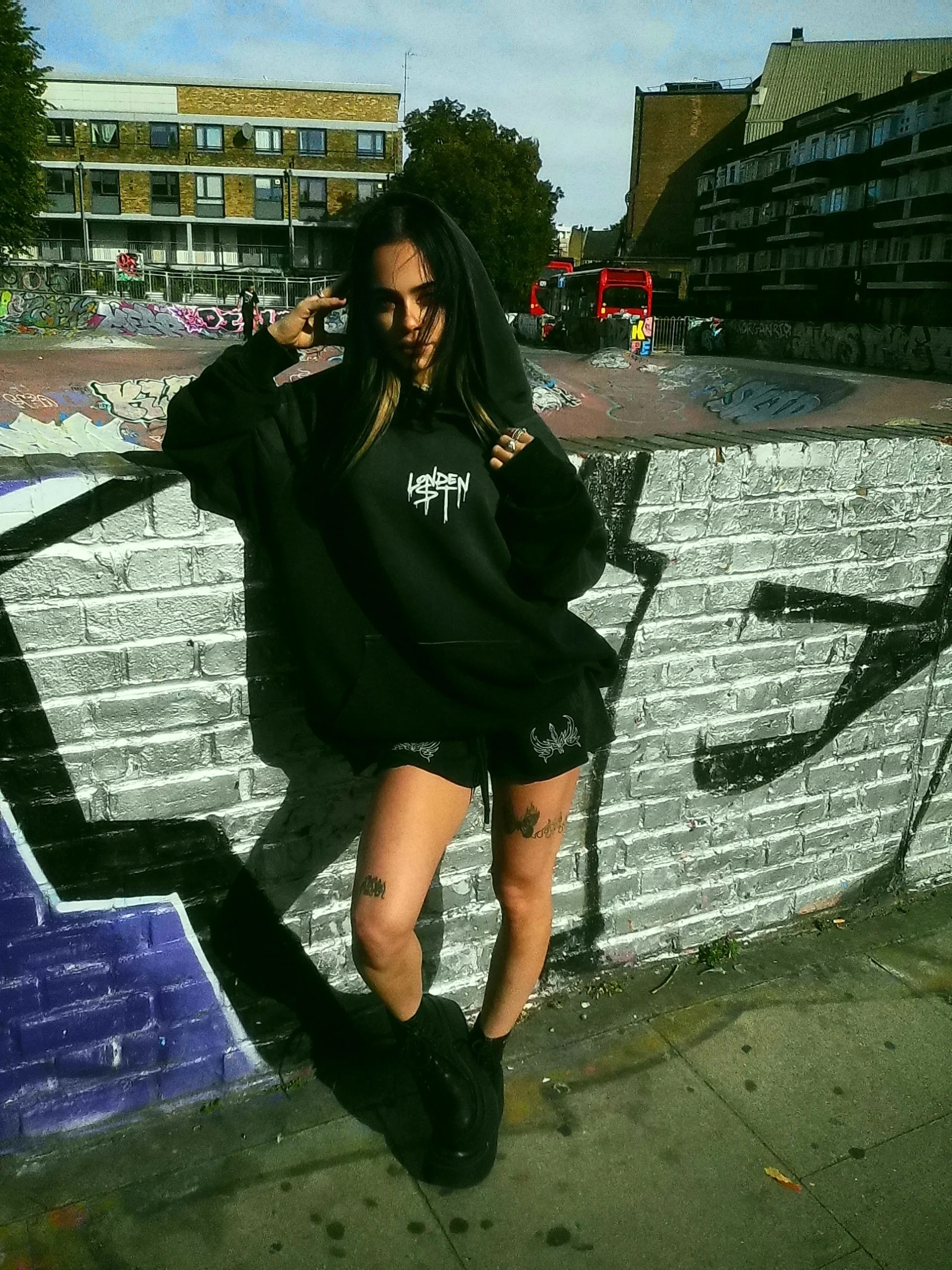 A young woman with long dark hair wearing a black hoodie, black shorts, and black boots standing against a graffiti-covered brick wall in an urban skate park with graffiti and residential buildings in the background.