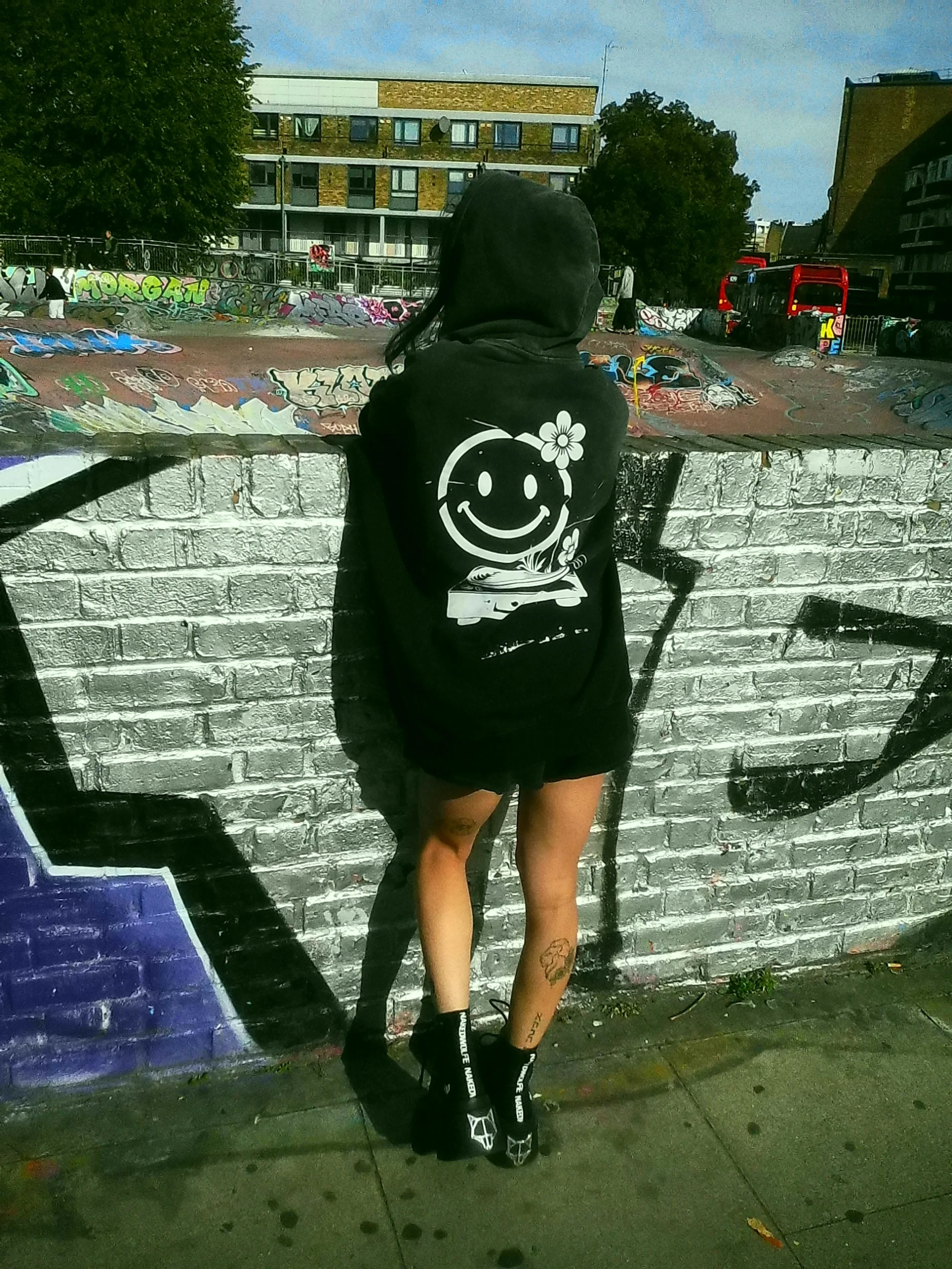 A woman in black hoodie with a smiley face graphic on the back, standing against a graffiti-covered brick wall in an urban park setting.