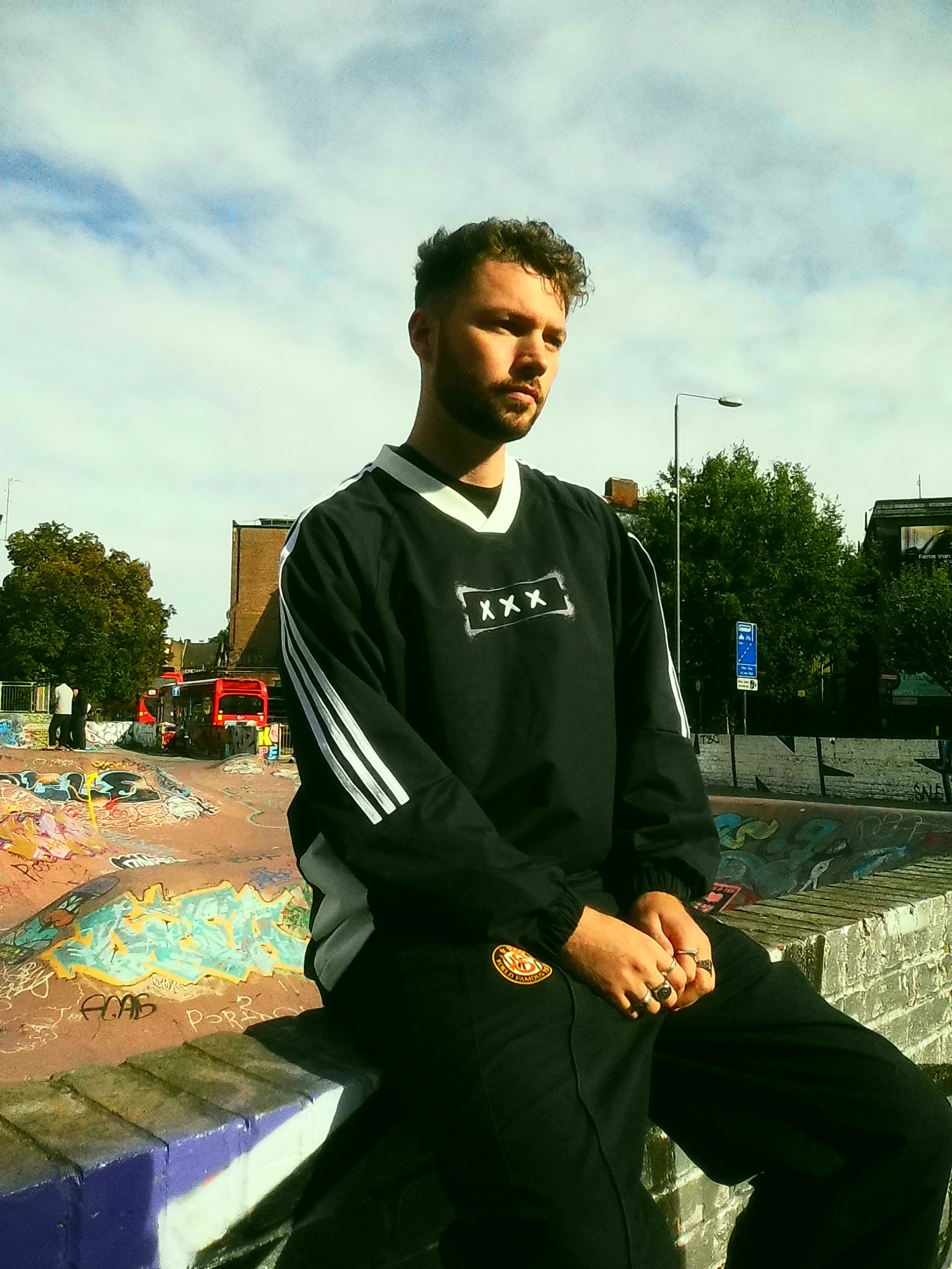 A young man with a beard and curly hair sitting on a graffiti-covered skatepark ledge, wearing a black sports jacket with white stripes and a black t-shirt. The background features graffiti art, trees, and a cloudy sky.