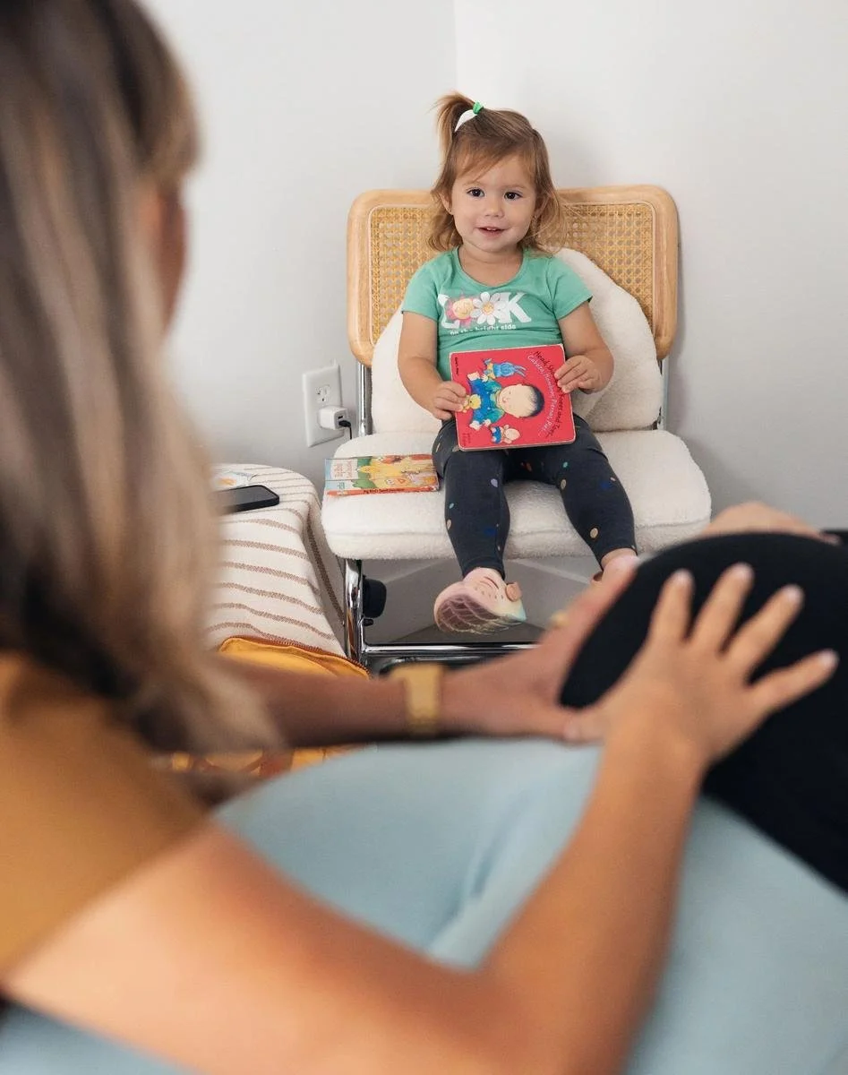 Smiling toddler in a green shirt on a cane chair while her pregnant mother receives a belly adjustment — pelvic pain and SPD chiropractic care in Solana Beach