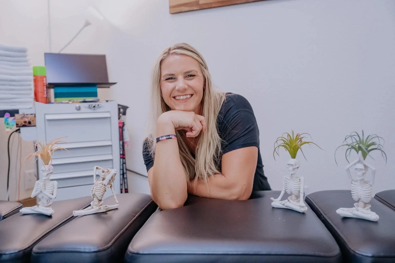 Dr. Jordan Brand in a black clinic top leaning on the adjustment table with anatomical skeleton models in the background — chiropractic care for Cardiff-by-the-Sea patients