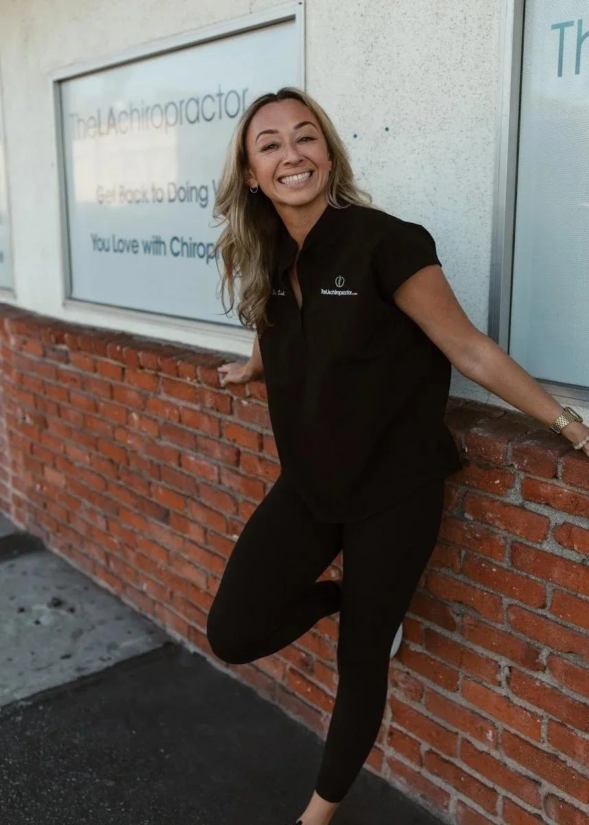 Dr. Lisel standing outside the practice in black scrubs, hand on hip, smiling against a brick wall and clinic signage — at Dr. Lisel Lewis Chiropractic in Solana Beach CA