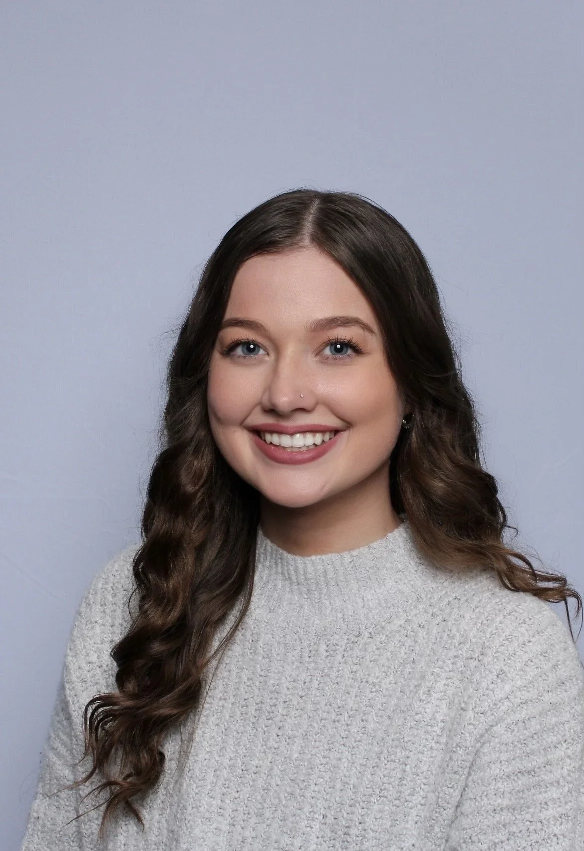 A young woman with long, wavy brown hair and blue eyes, smiling broadly, wearing a light gray sweater, against a plain light gray background.