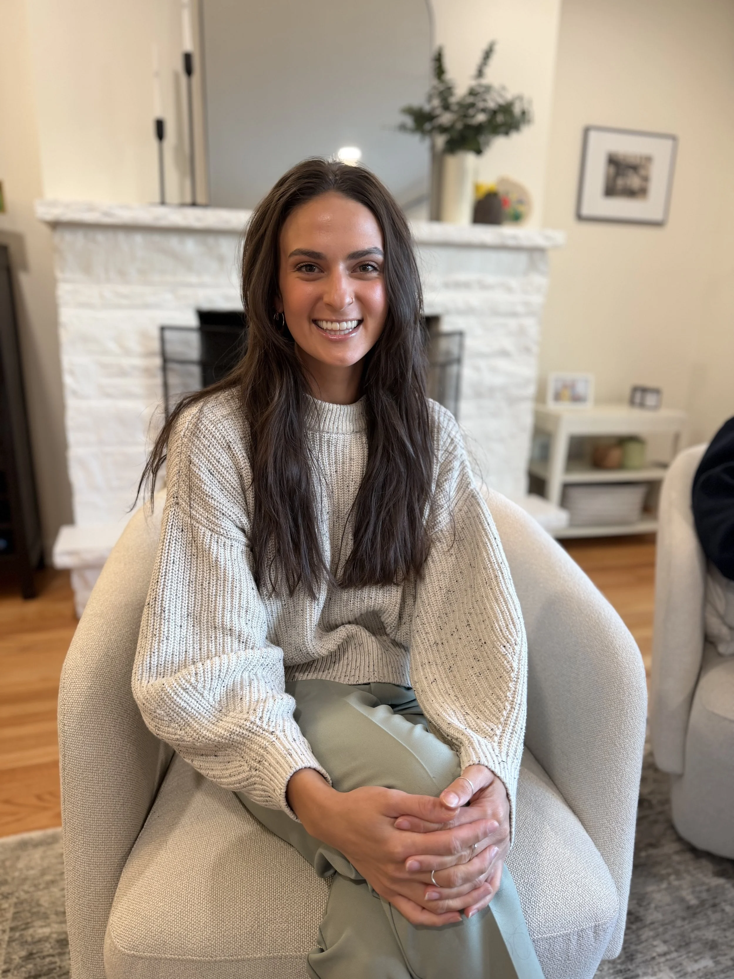 A smiling woman with long dark hair sitting on a beige armchair in a cozy living room, with a fireplace and a potted plant in the background.
