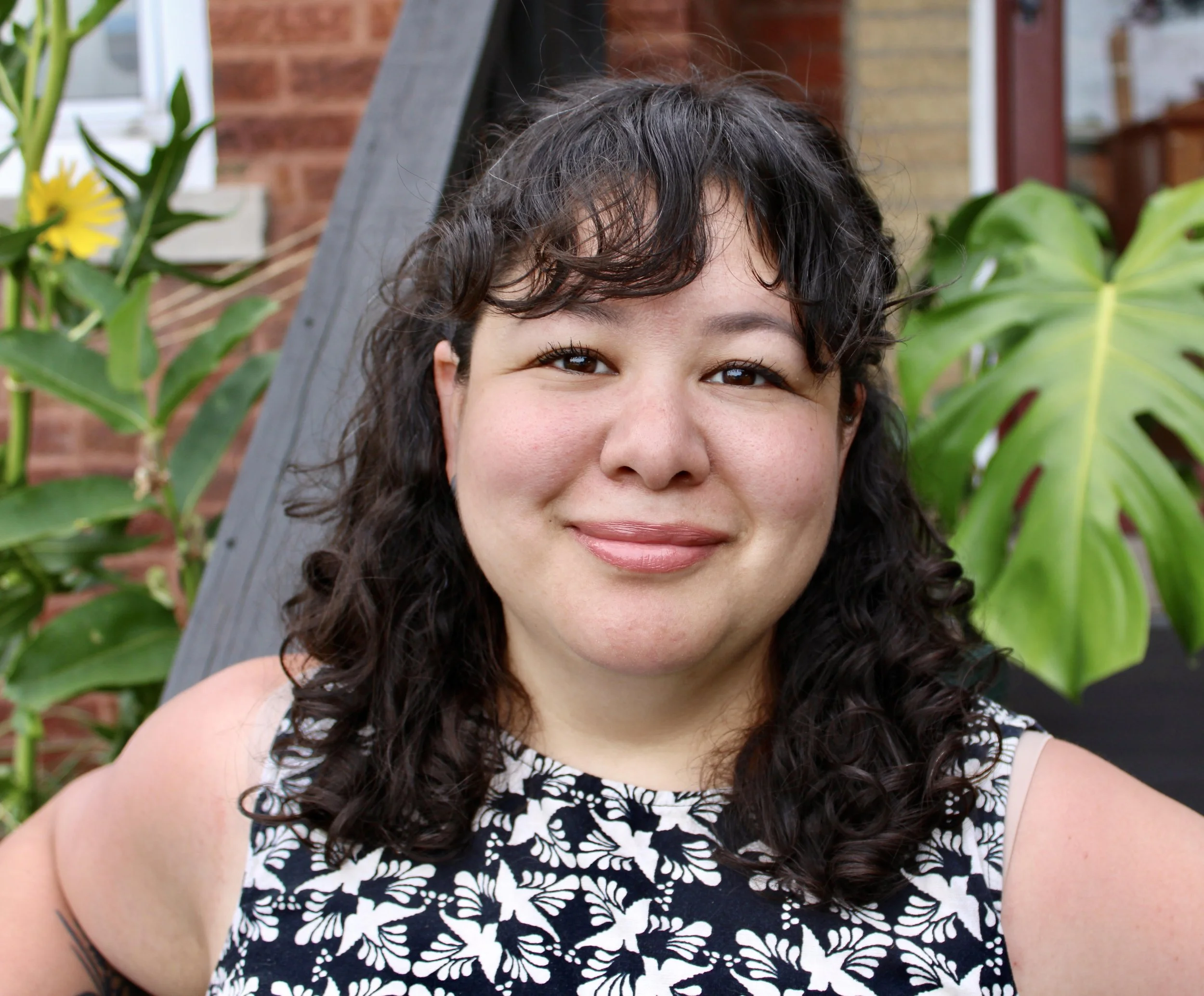 Close-up of a smiling woman with long, curly dark hair and makeup, wearing a white and black patterned top, standing outdoors with green plants and a brick wall in the background.
