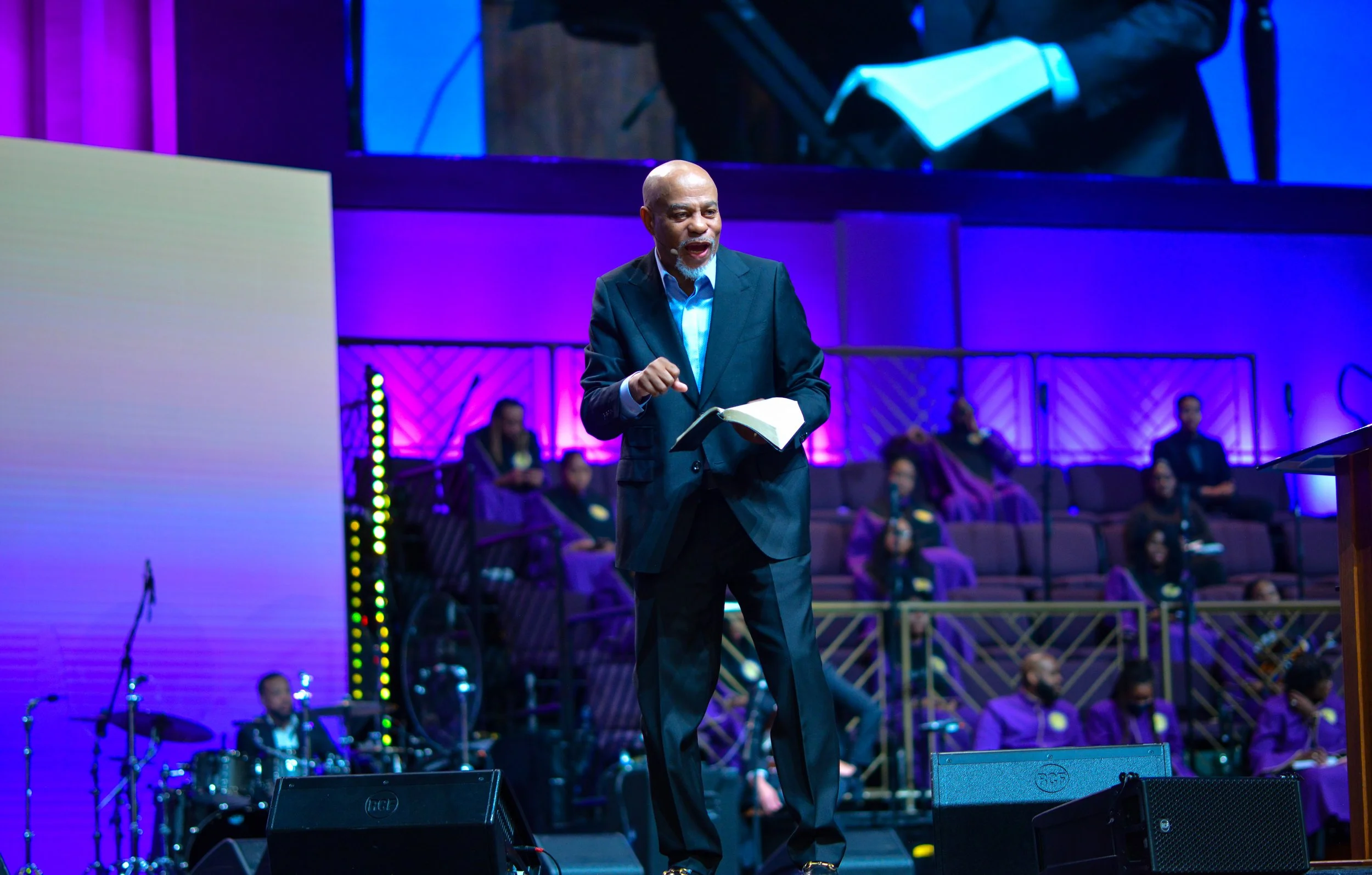 A man in a dark suit speaking at a podium during a church service, with a choir and musicians in purple robes in the background.