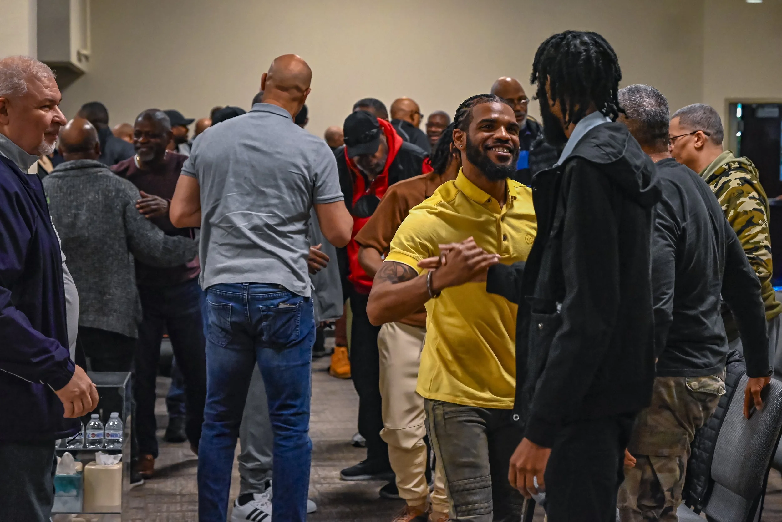 A group of people gathered indoors, where two men are shaking hands and smiling in the foreground, with others socializing in the background.