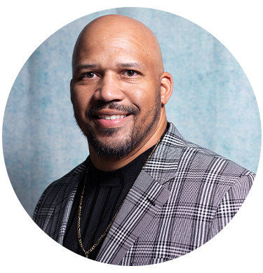Portrait of a smiling man with a bald head and a neatly trimmed beard, wearing a plaid blazer and a black shirt, against a light blue background.