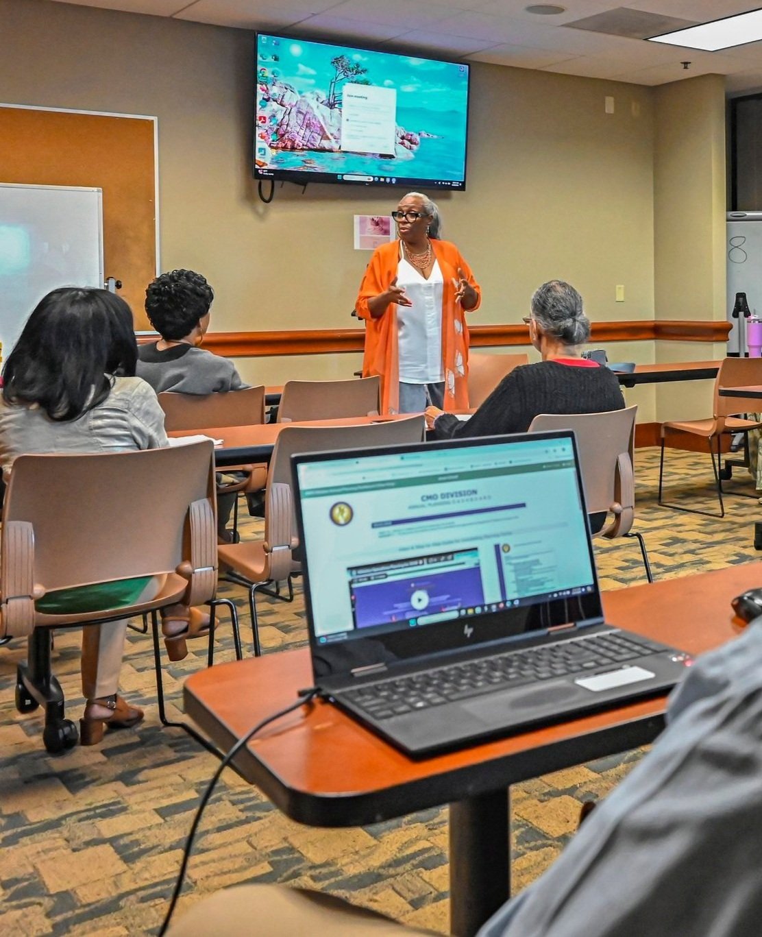 A woman giving a presentation in a classroom or conference room to a small group of adult participants, with a large screen behind her and a laptop on a desk in the foreground displaying a webpage.