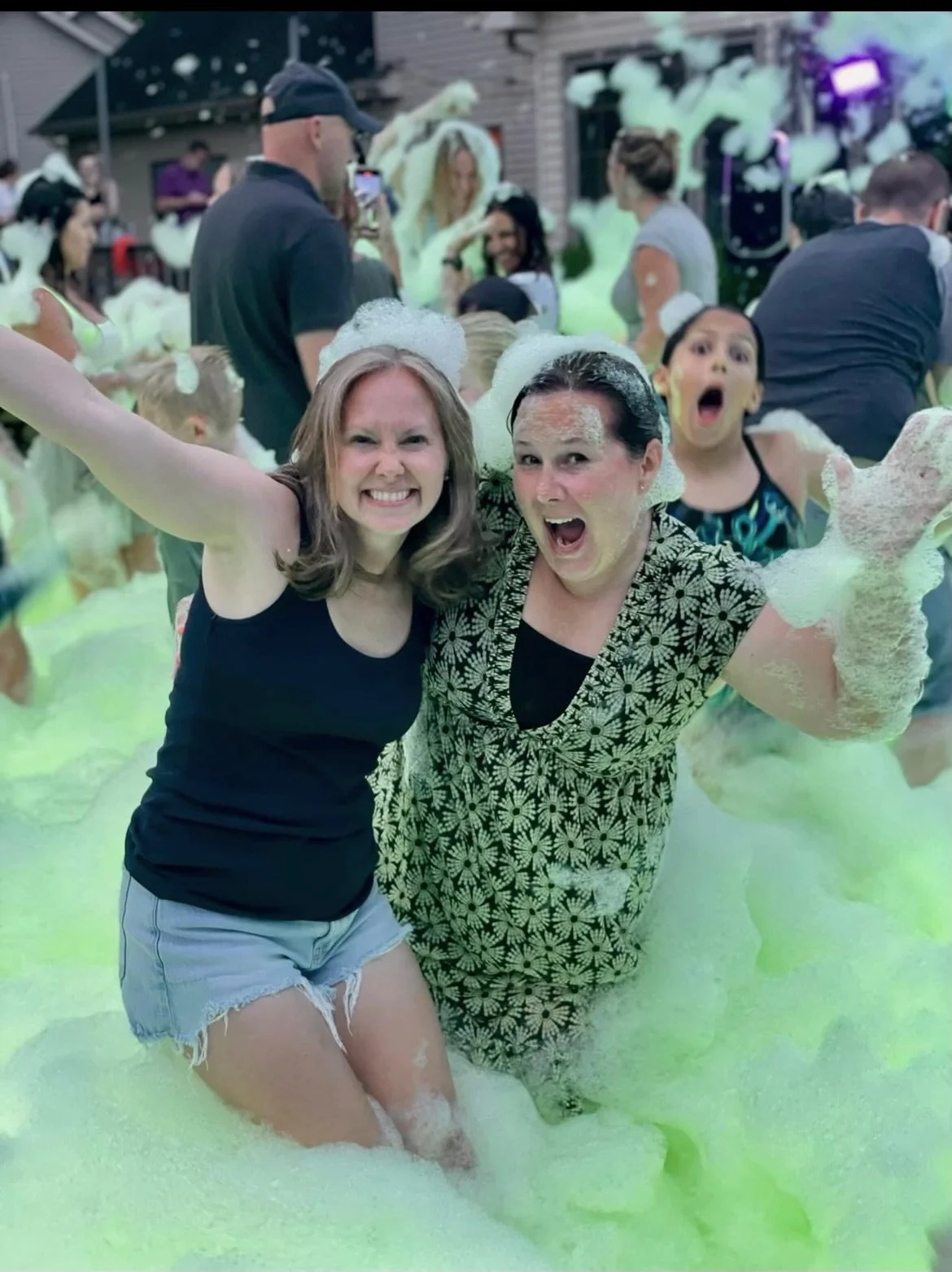 Two smiling women enjoying a Varsity Foam adult foam party in Chicagoland surrounded by green foam at an outdoor event