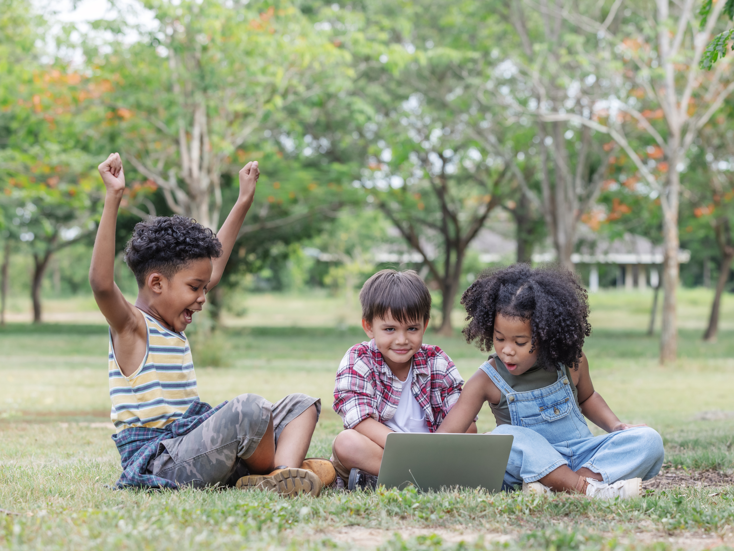 three young kids sitting on the grass outside and using a laptop