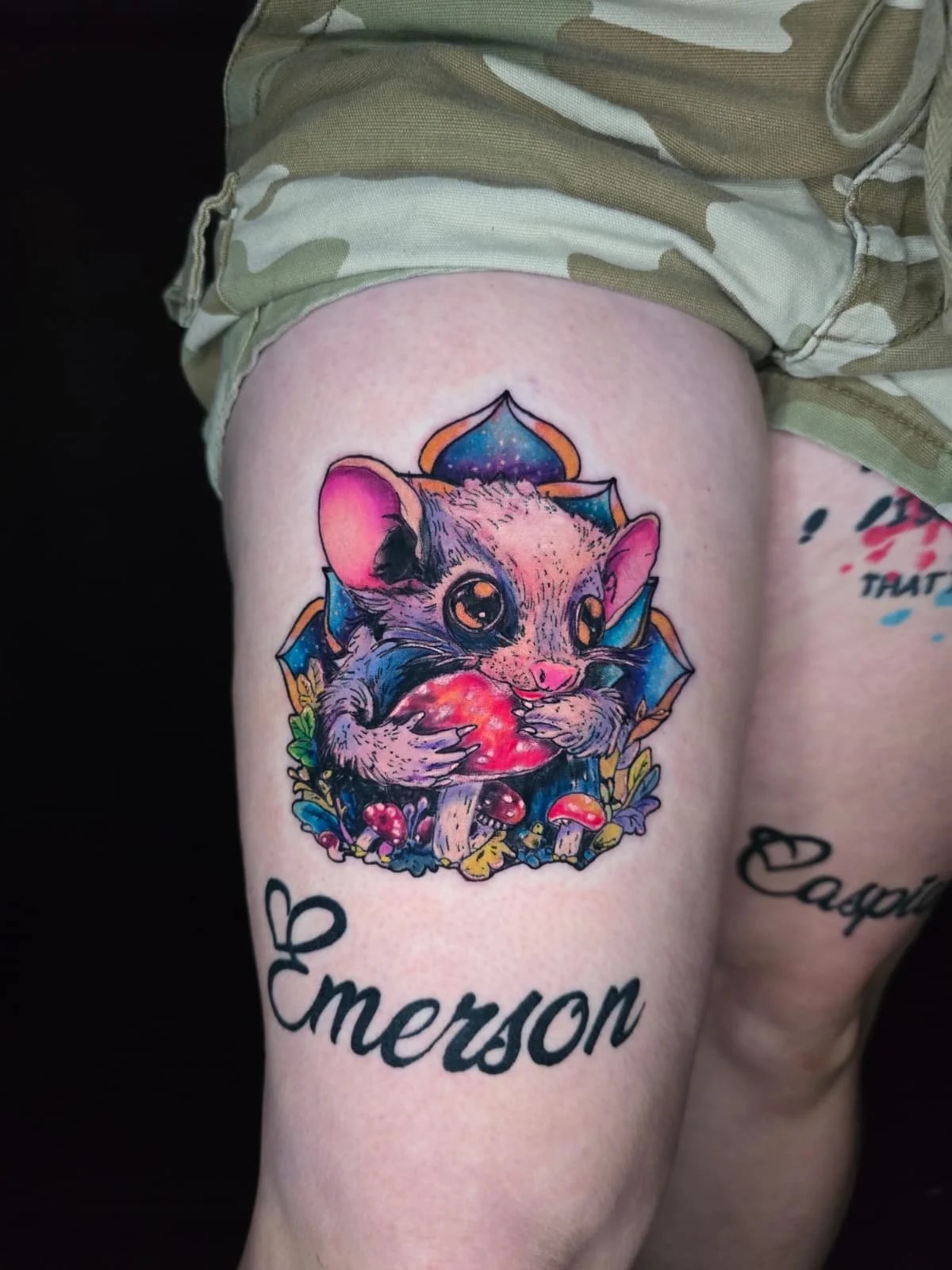 Colorful tattoo of a cute mouse holding a red mushroom, surrounded by smaller mushrooms and leaves, with the name 'Emerson' written in cursive below.