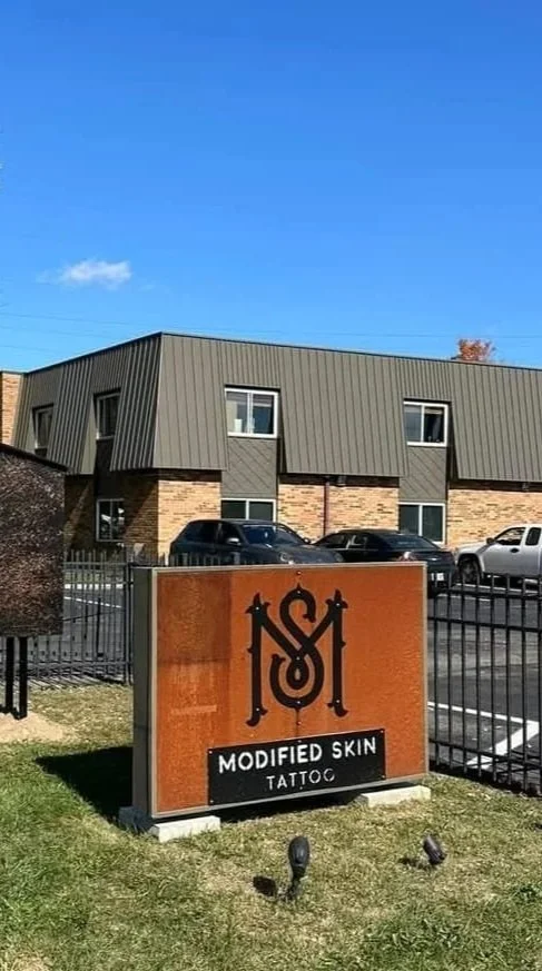 A sign for a tattoo shop called Modified Skin Tattoo in front of a parking lot, with a modern two-story building and parked cars in the background against a blue sky.