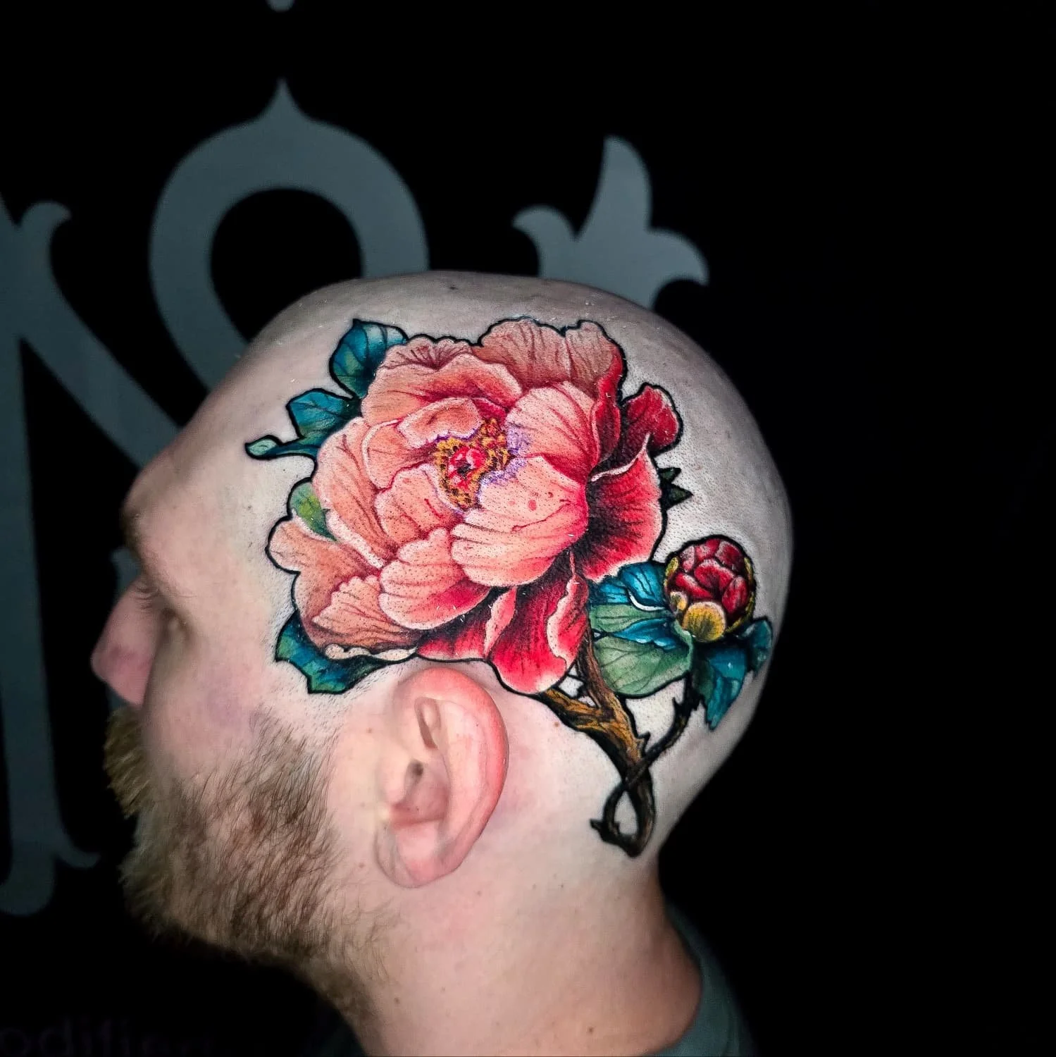 Side profile of a person with a large, colorful tattoo of a pink peony flower and buds on their head and scalp, with visible beard and ear.