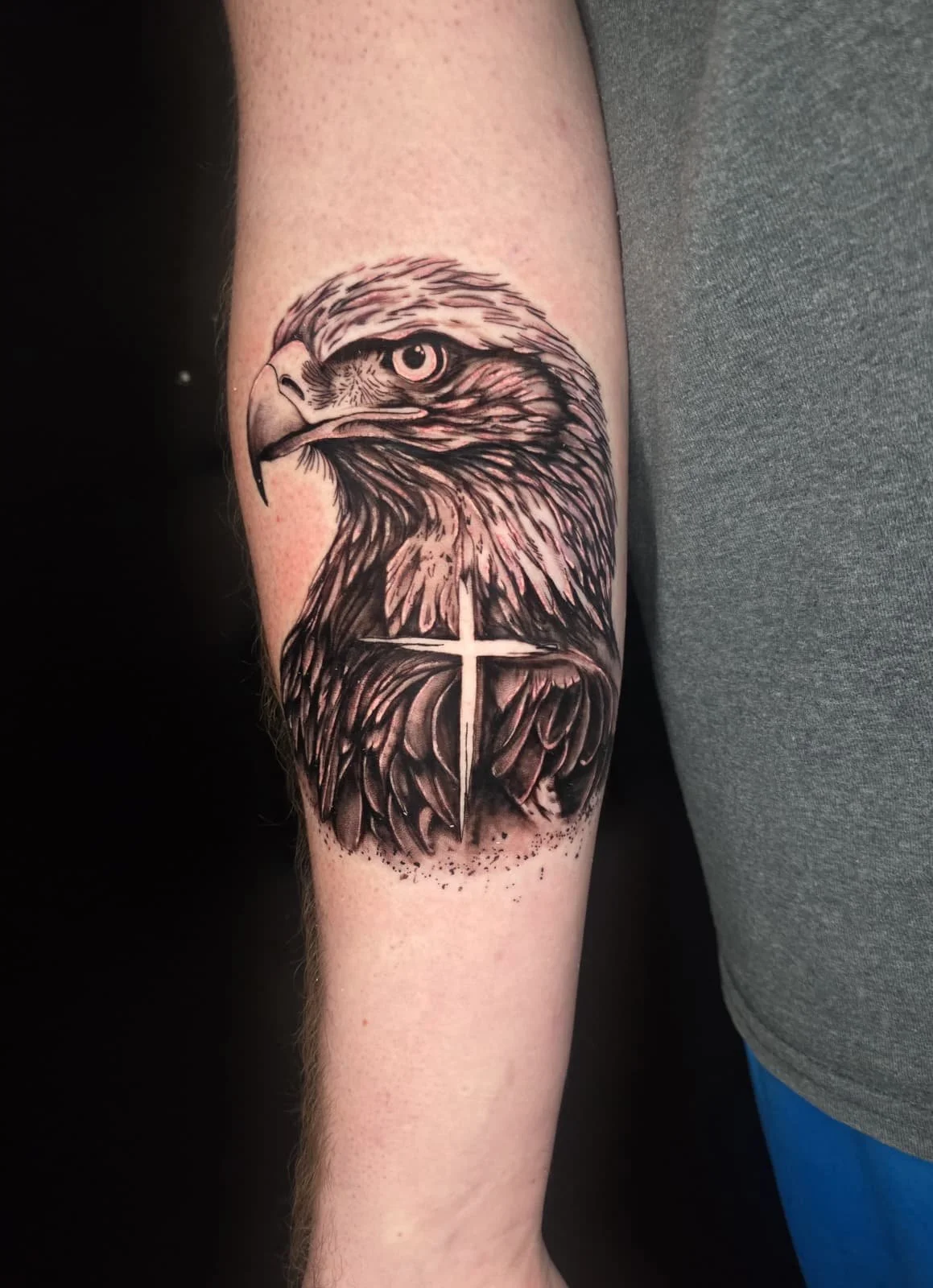 Black and gray tattoo of an eagle's head with detailed feathers and an intense eye, featuring a white cross integrated into the eagle's feathers.