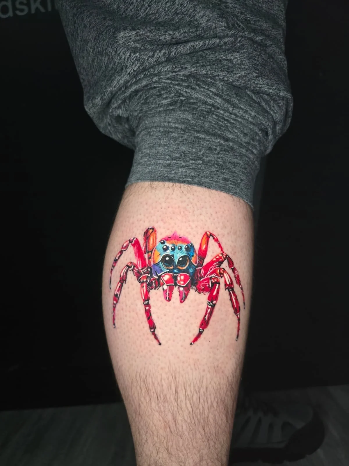 Colorful tattoo of a spider with a cartoonish face on a person's leg.