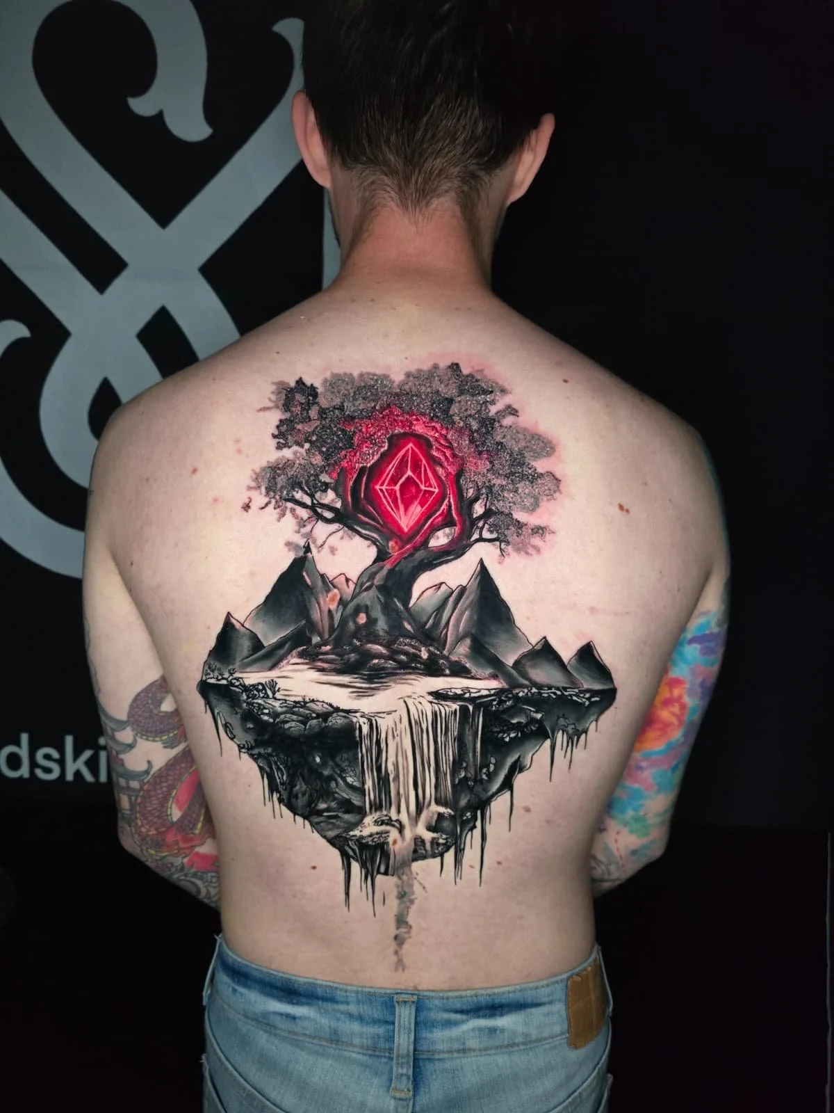 A person with tattoos on both arms displaying a large tattoo on their back of a landscape with mountains, trees, a waterfall, and a glowing red geometric shape in the sky.