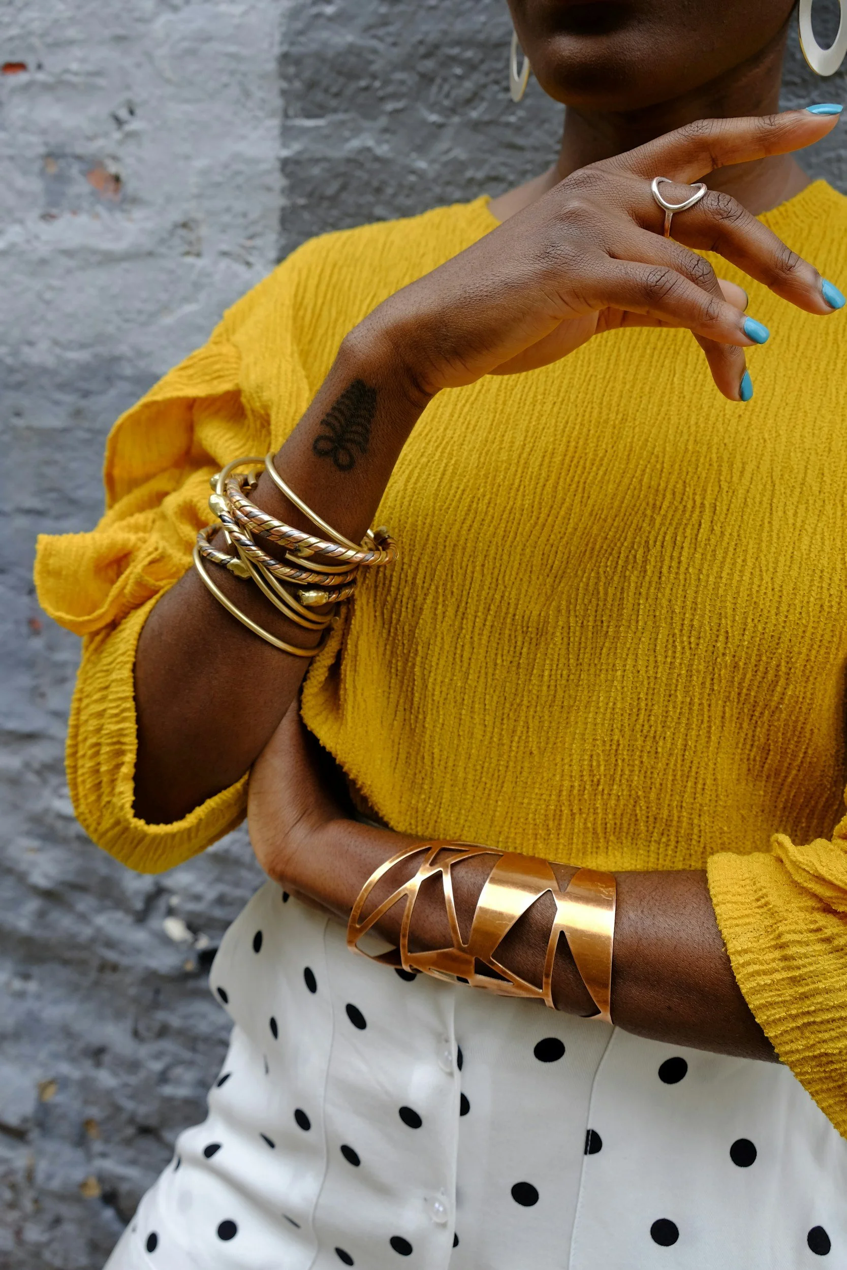 A woman wearing a bright yellow textured top, white polka dot skirt, and multiple gold bracelets on her wrist. She has a tattoo on her hand, turquoise nail polish, and large earrings, standing against a gray brick wall.