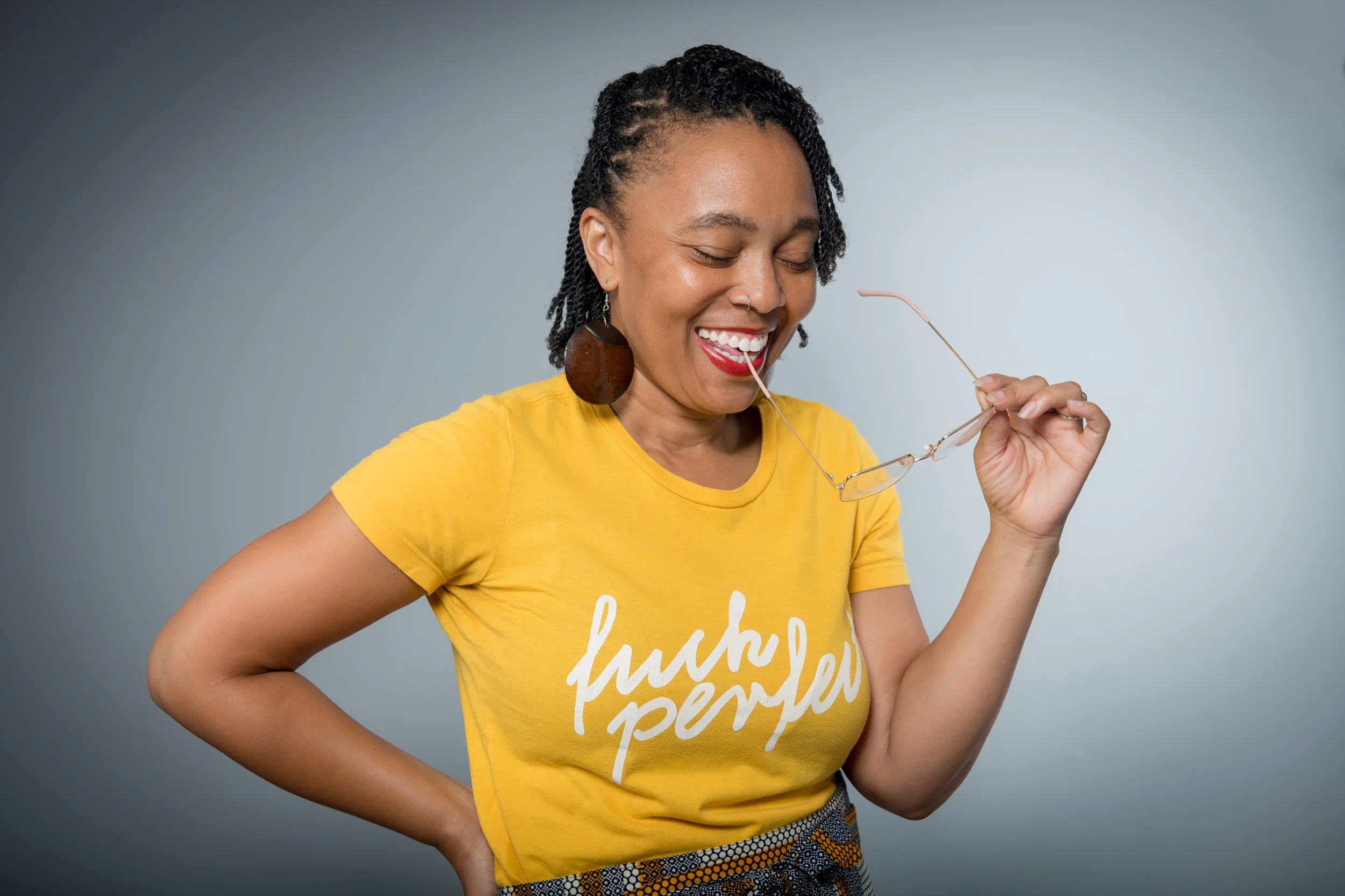 A woman with short, textured hair wearing a yellow T-shirt with white text and holding a pair of glasses. She is smiling and looking down against a neutral gray background.