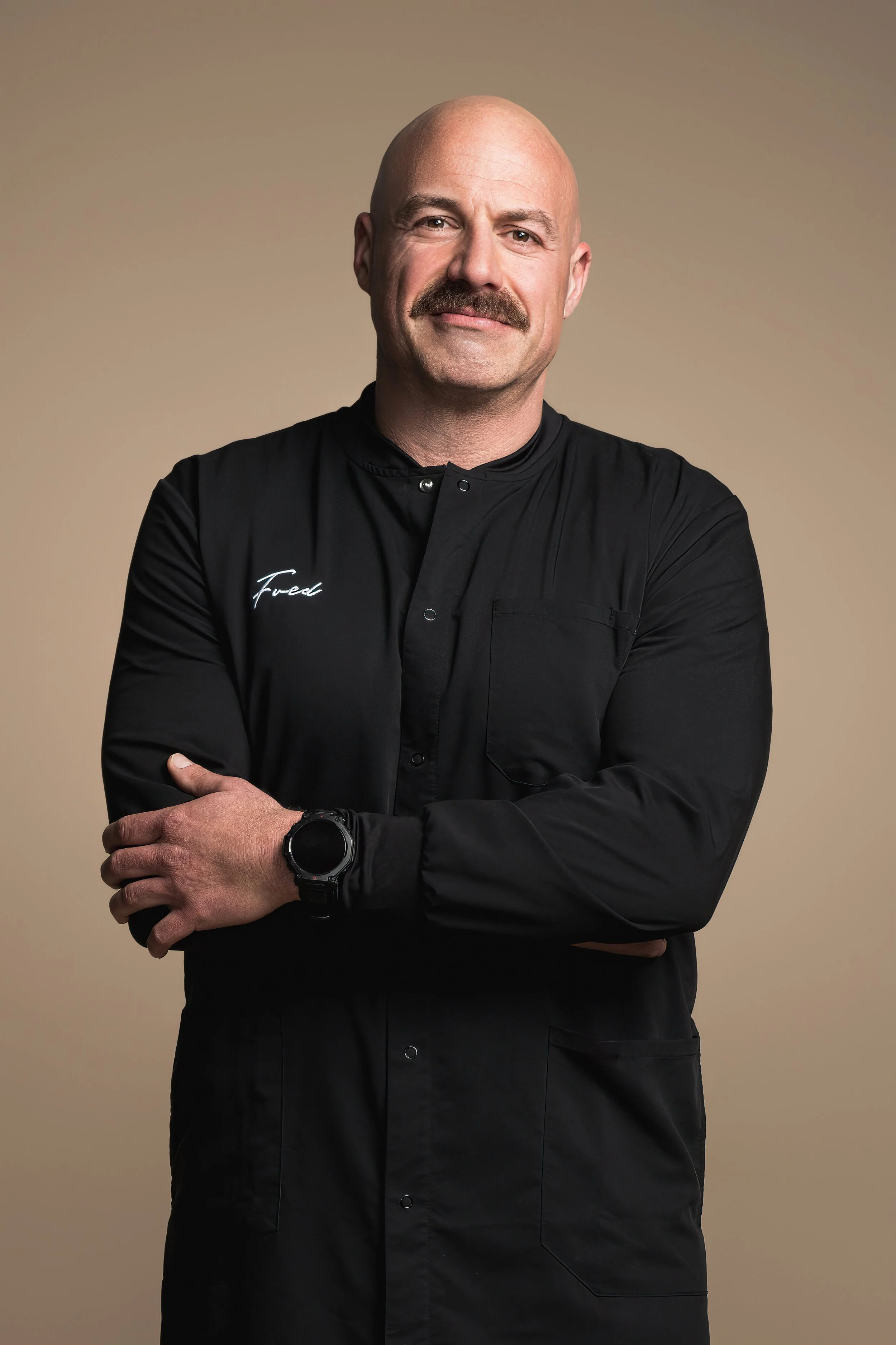 A man with a shaved head and mustache standing with arms crossed, wearing a black chef's jacket with the word 'Fused' embroidered on it, against a plain beige background.
