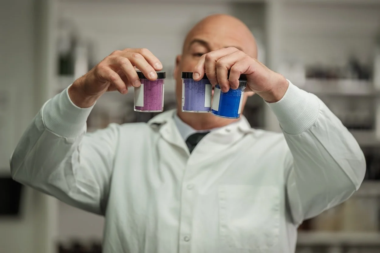 Man in a lab coat holding three containers of colored powder: pink, purple, and blue.