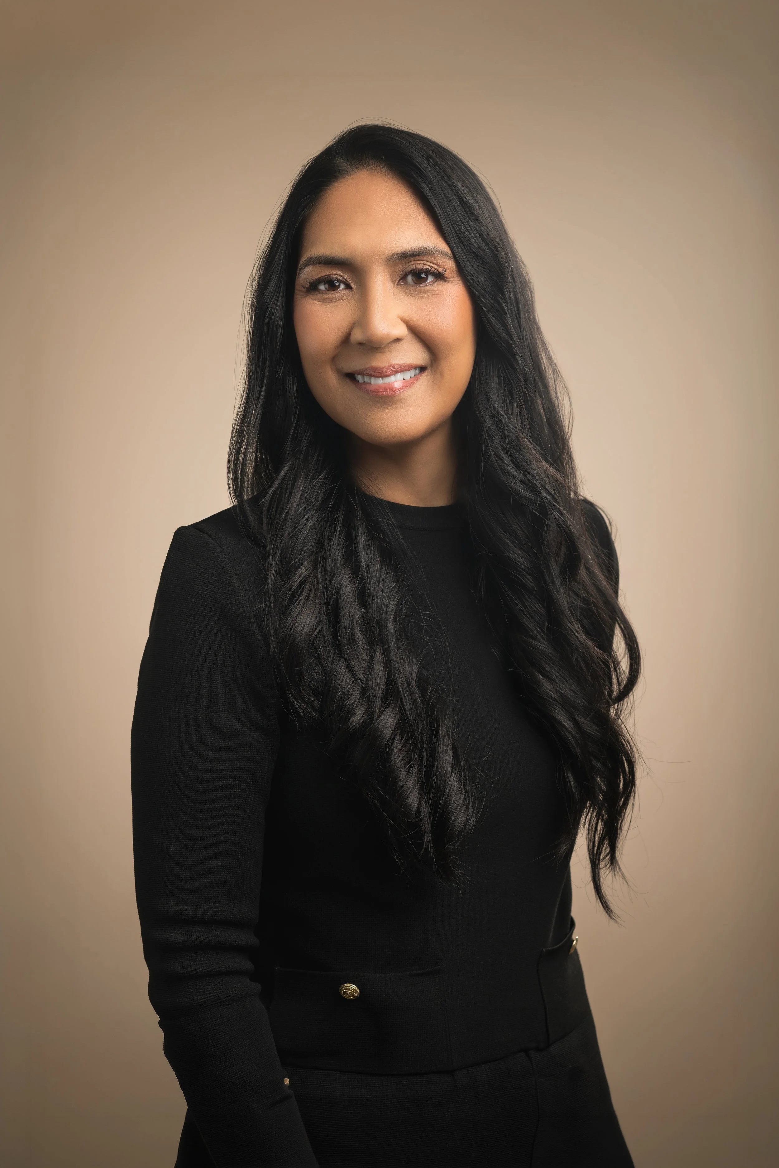 A professional woman with long black hair, wearing a black blazer with gold buttons, smiling against a plain beige background.