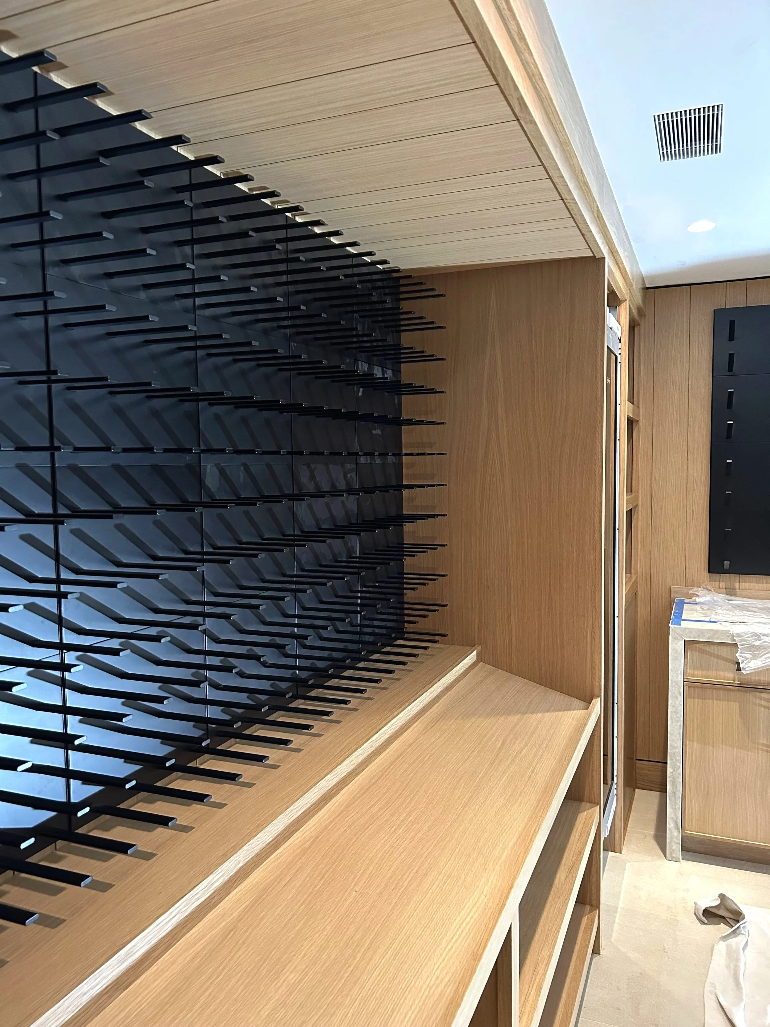 Interior view of a modern kitchen or bar area with a black wine rack, wooden cabinetry, and a countertop, with some construction or renovation materials visible.