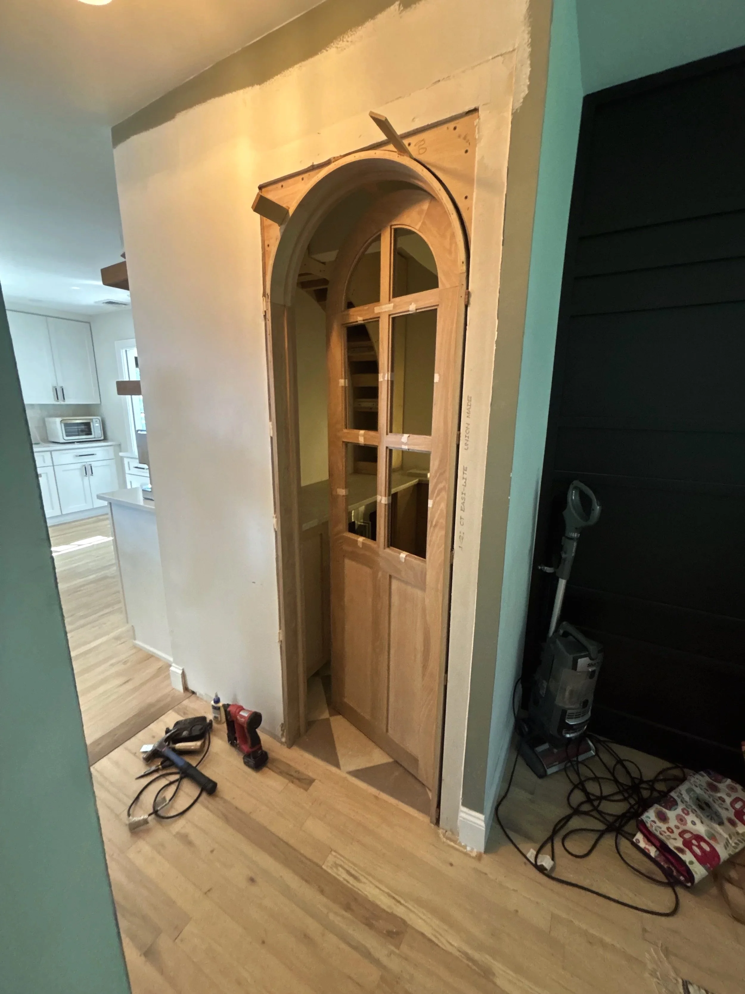 Interior view of a partially installed wooden door with an arched top and muntin windows, next to a black wall and a vacuum cleaner, with construction tools on the wooden floor.