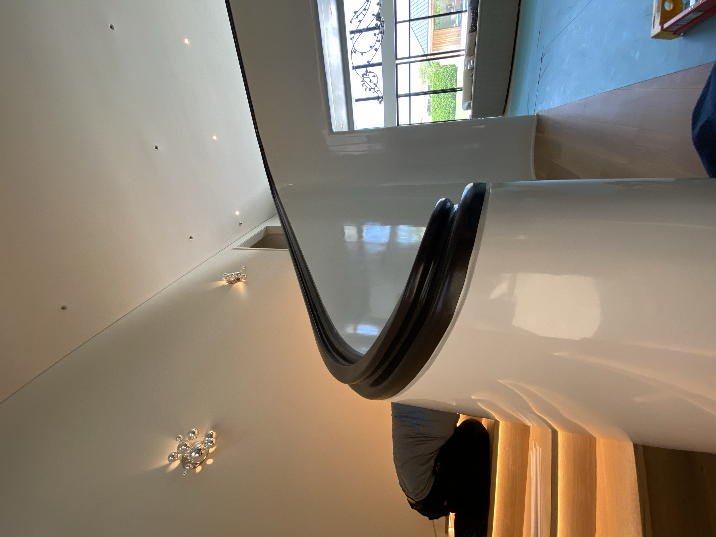 A white, rounded countertop with a black trim edge in a modern interior space. In the background, there is a large window, some furniture, and greenery outside, with a person's arm and part of their torso visible.