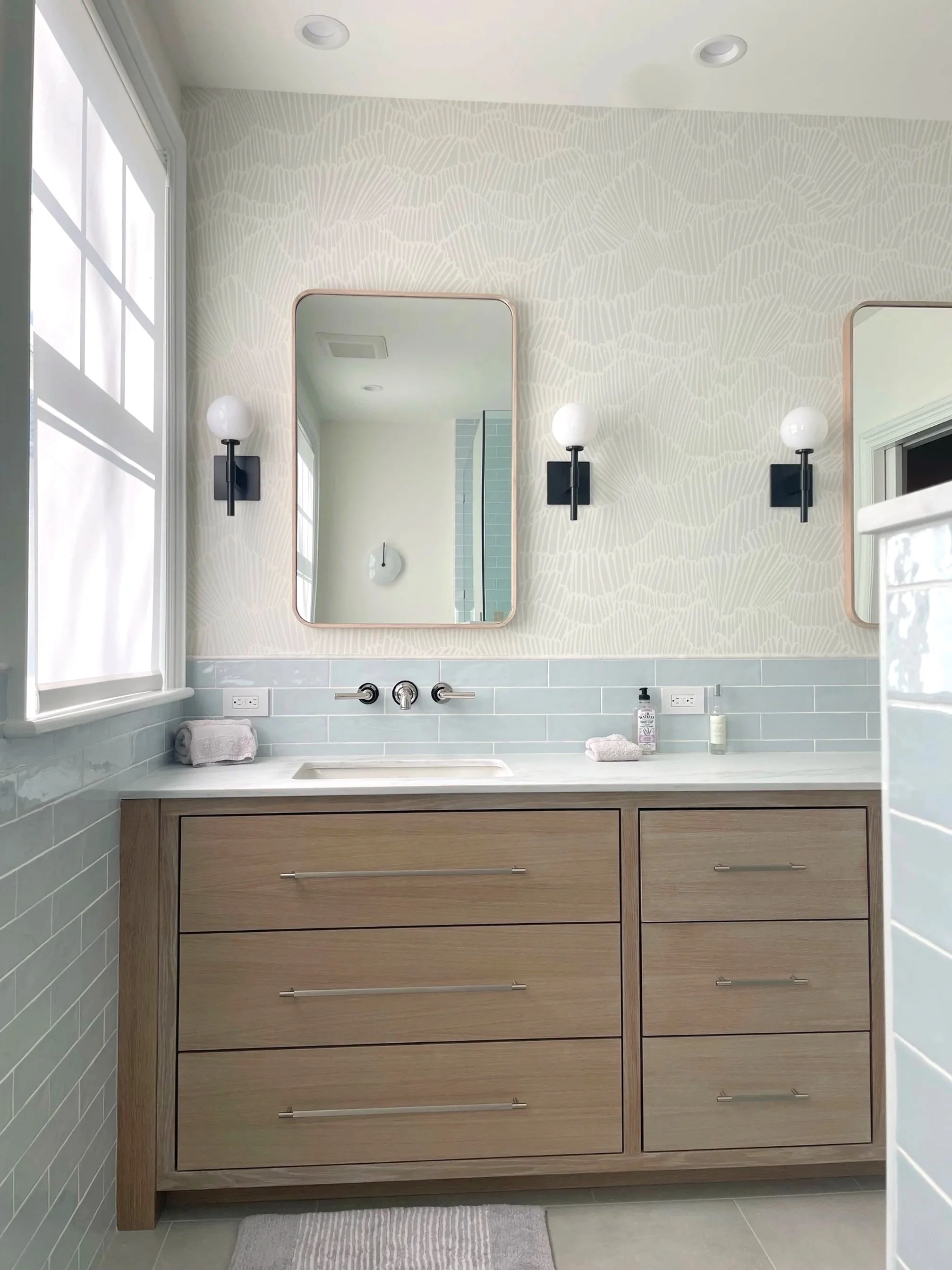 A modern bathroom vanity with two mirrors and black wall sconces, a light-colored wooden cabinet, white countertop, and a bathroom window with white blinds.