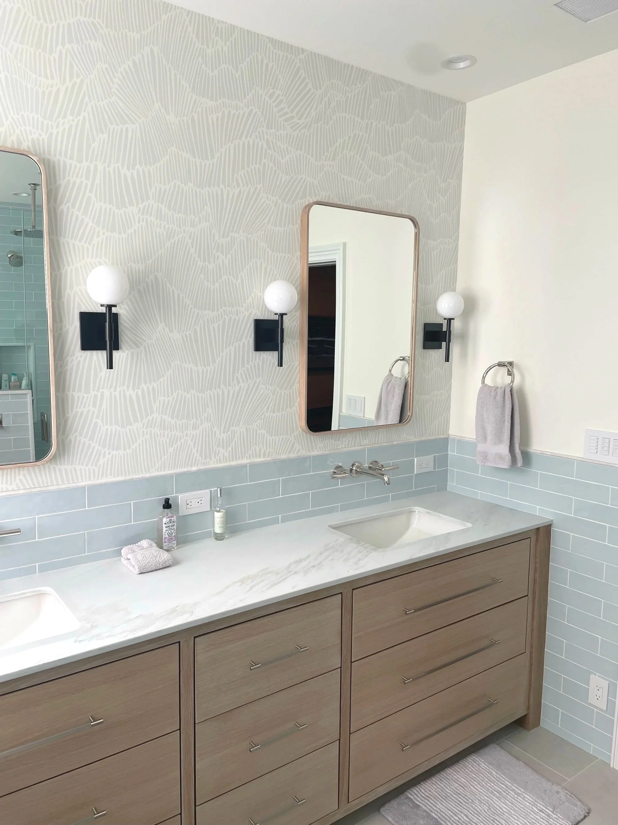 Modern bathroom with light wood vanity, two mirrors, and a patterned wallpaper accent wall. Two black sconces with globe lights flank each mirror.  Custom vanity renovation in The Hamptons.