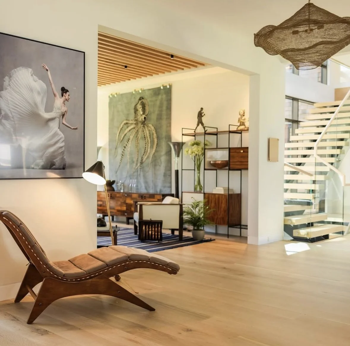 A modern living room with wooden floors, a chaise lounge, and a large black and white wall art of a ballet dancer. In the background, there are decorative sculptures, plants, and a staircase with glass railing and wooden steps.