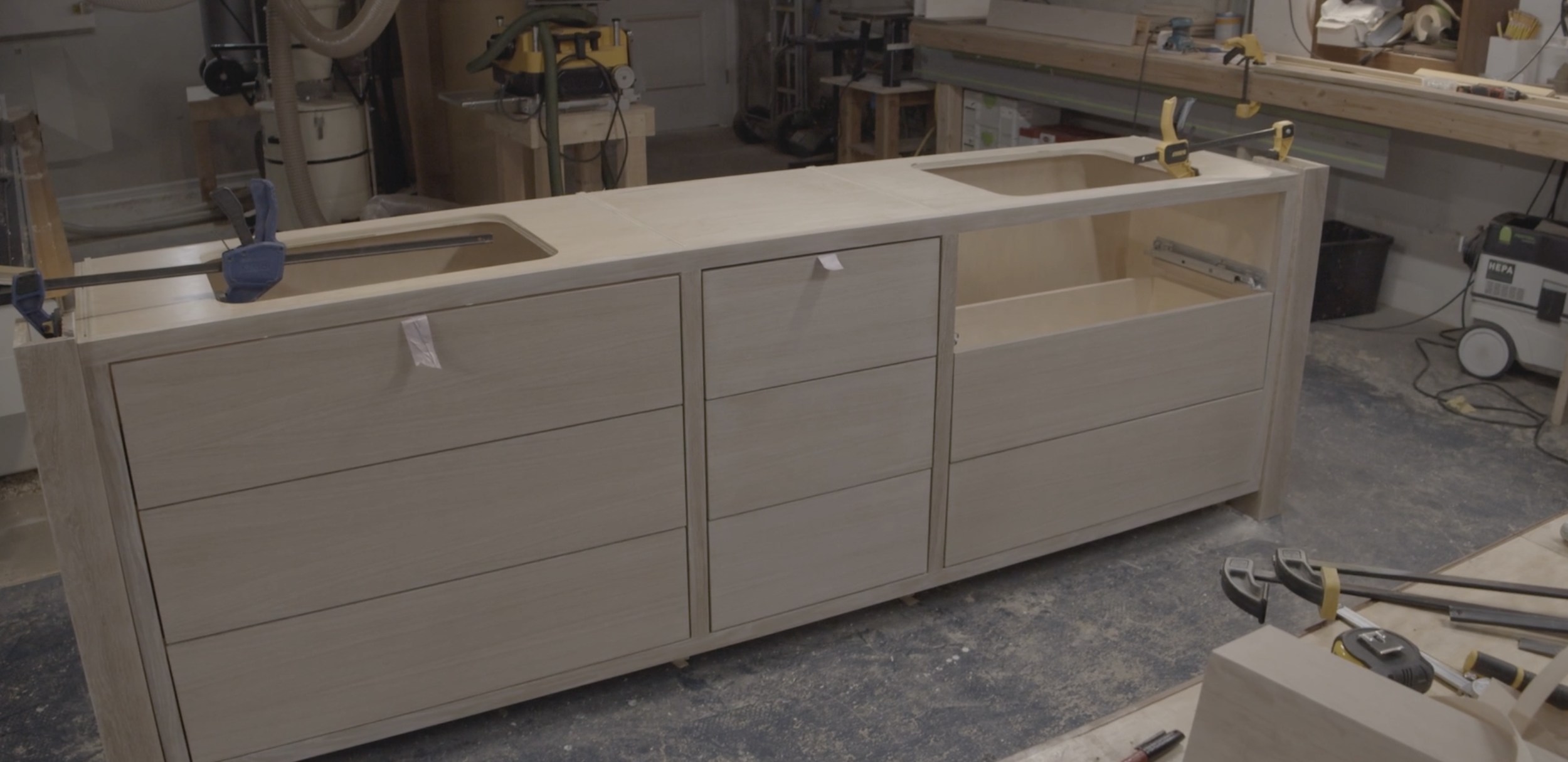 Wooden kitchen cabinet under construction in a woodworking workshop, with clamps holding parts together and tools nearby.