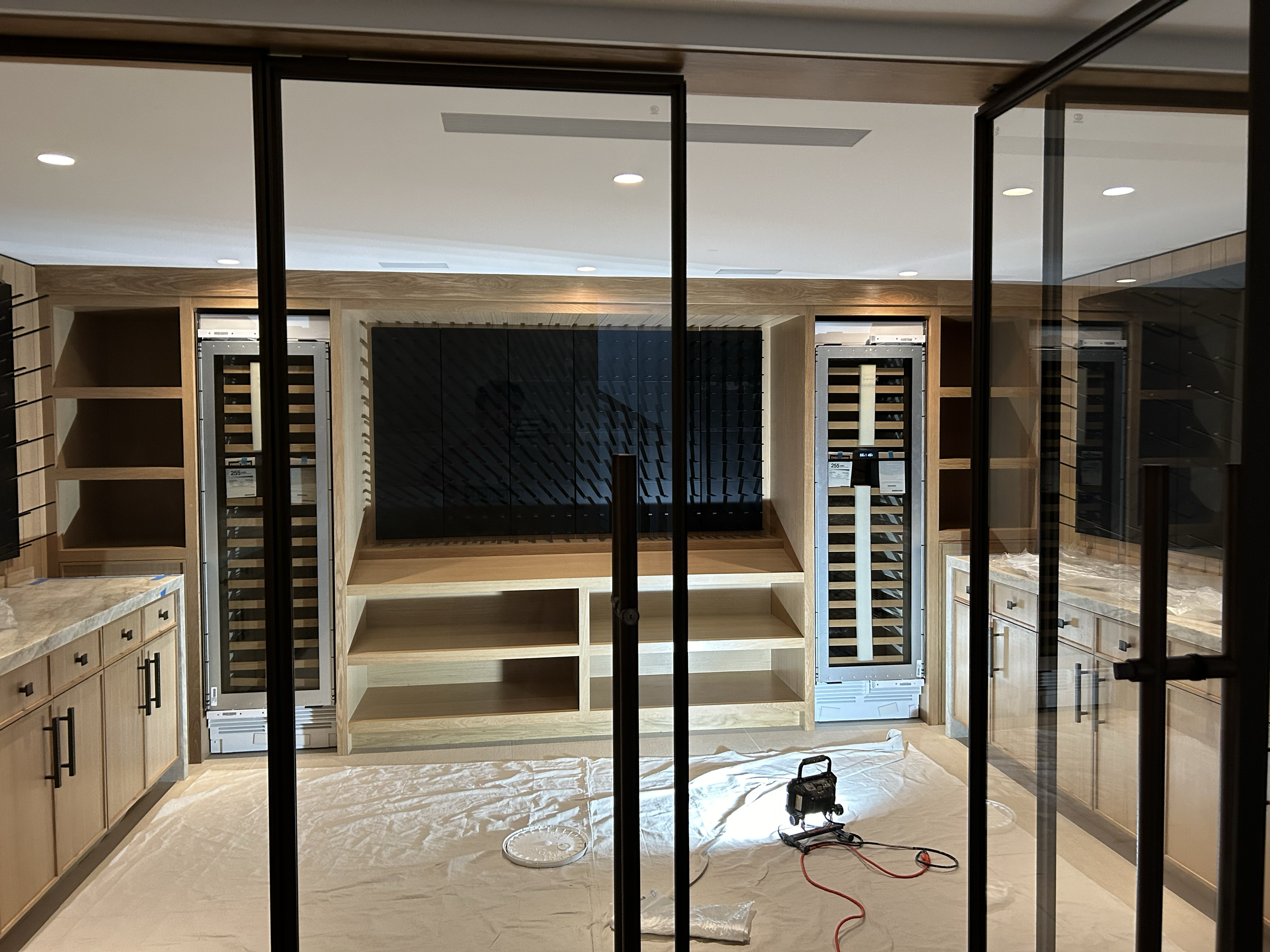 View of a kitchen or entertainment room under construction, with wood cabinetry, open shelving, and two wine refrigerators, seen through a glass wall or doors.