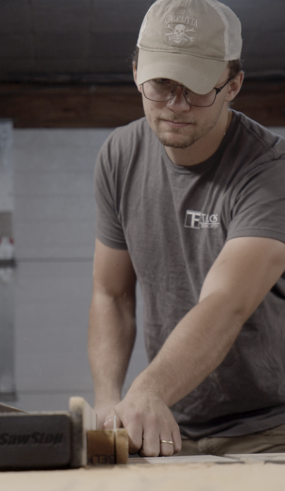 A young man wearing glasses, a beige cap, a gray t-shirt, and a wedding ring, working with a saw on a wooden piece in a workshop.