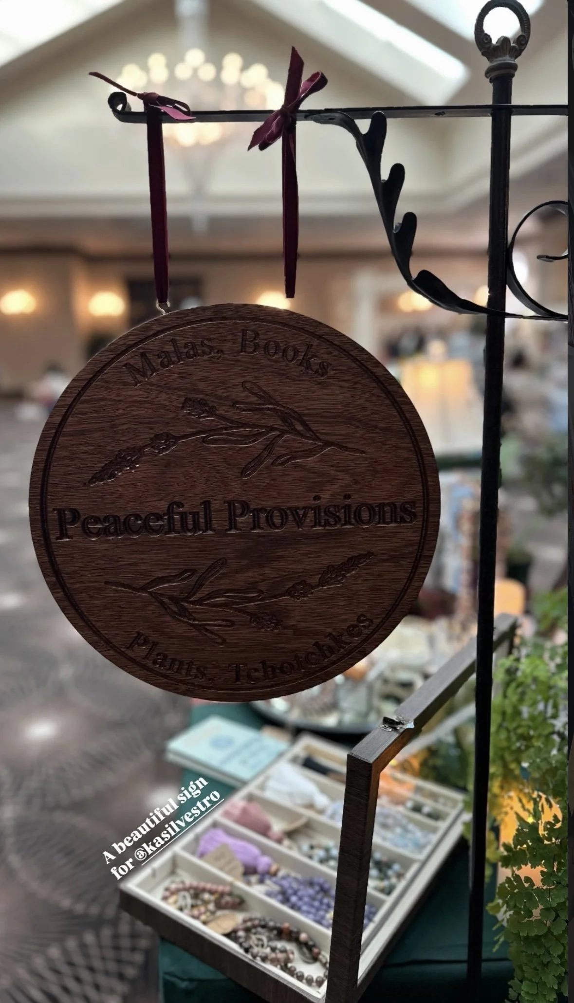Wooden round sign hanging from a black metal stand that reads "Madas Bokes" at the top and "Peaceful Provisions" in the middle, with decorative branches engraved. Below the sign, there's a display tray with jewelry and a card that says "A beautiful s