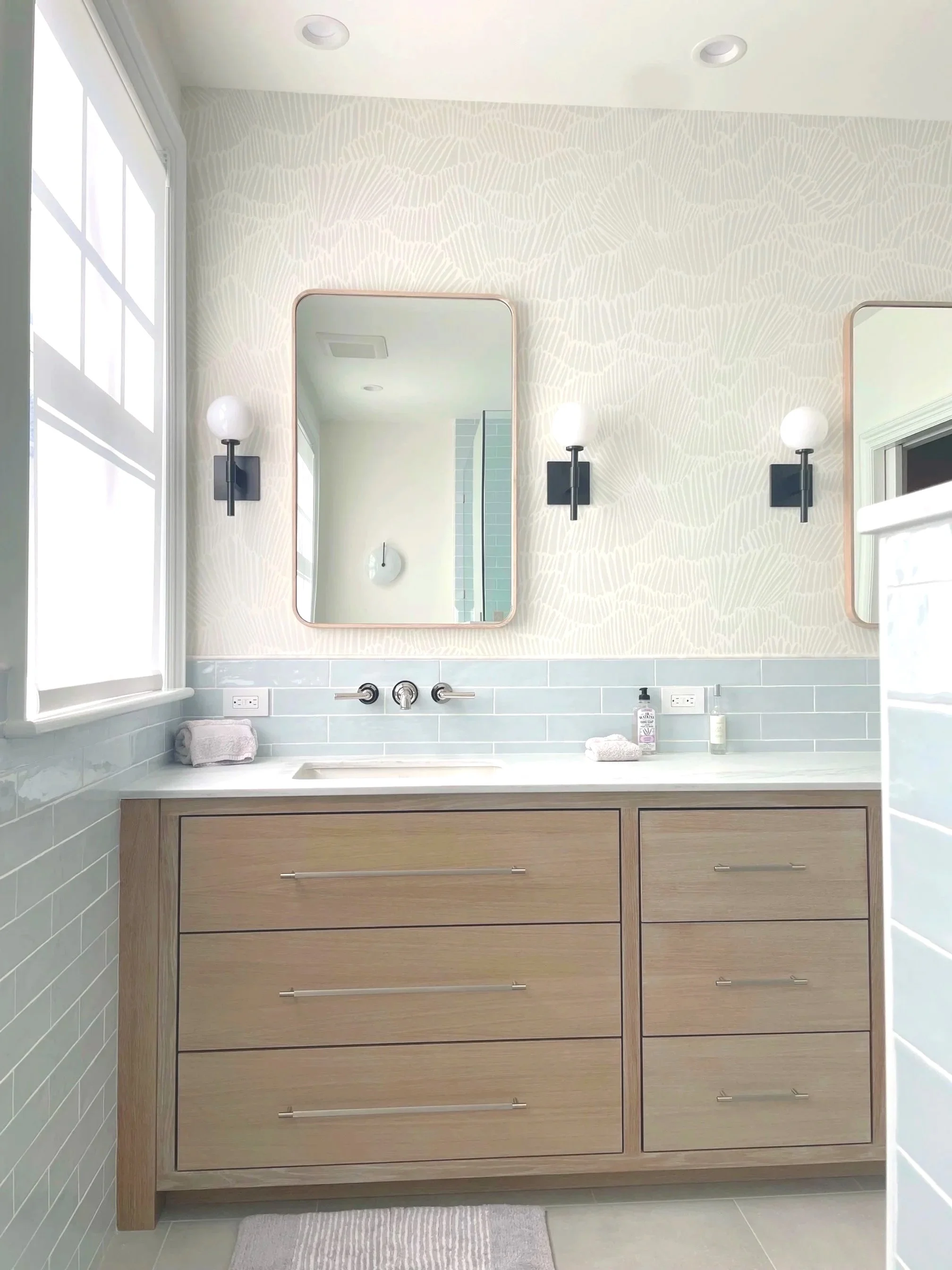 Modern bathroom with a wooden vanity, two mirrors, wall-mounted light fixtures, a window, and bathroom supplies on the countertop.
