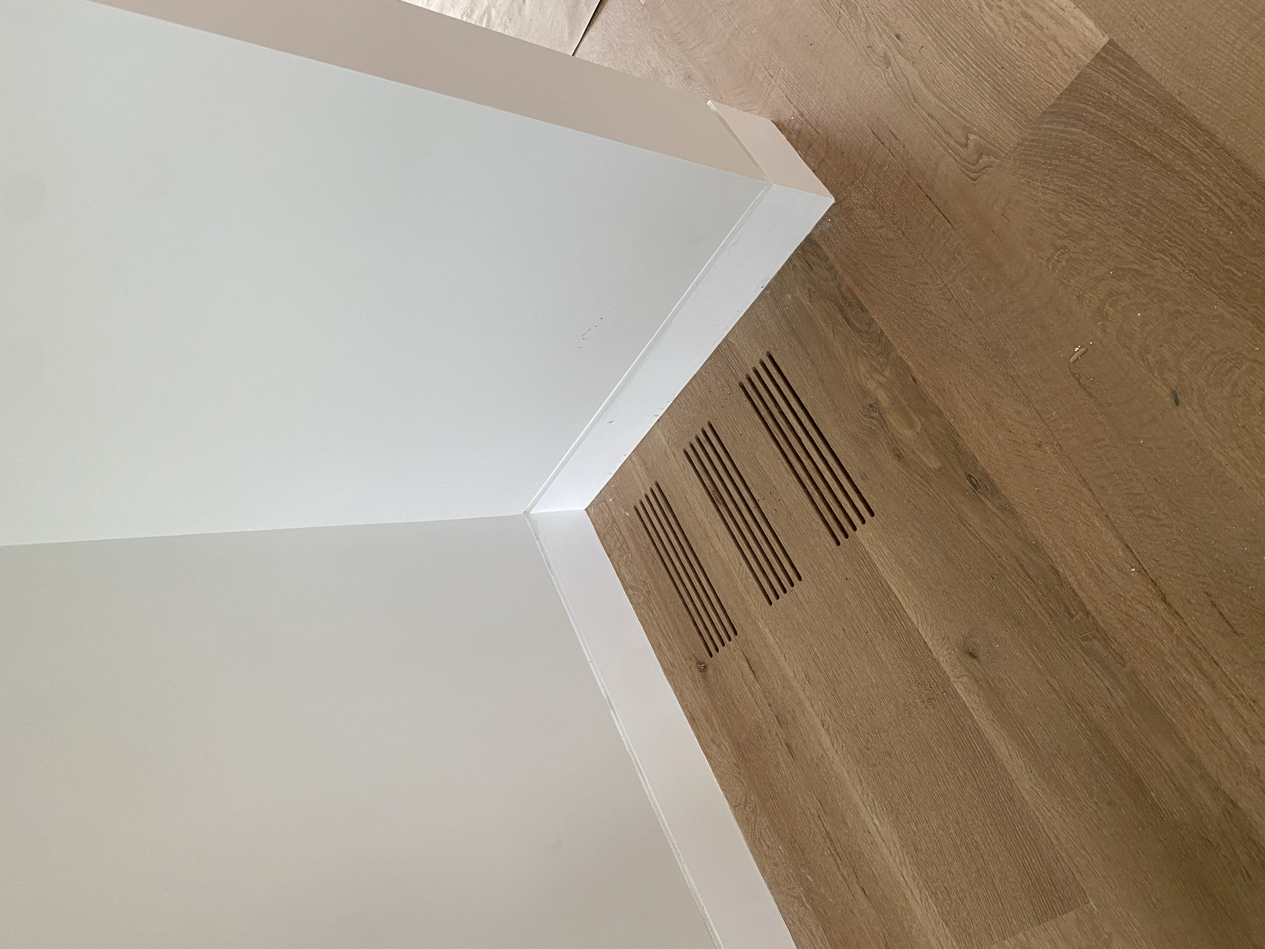 Corner of a room with a white wall, white baseboard, and brown wooden floor with air vent vents. AC Register.