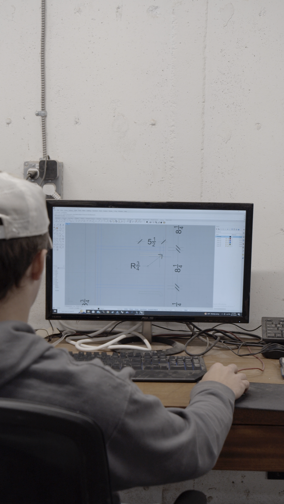 A person working at a desk on a computer with a schematic drawing on the screen, showing measurements and geometric shapes.