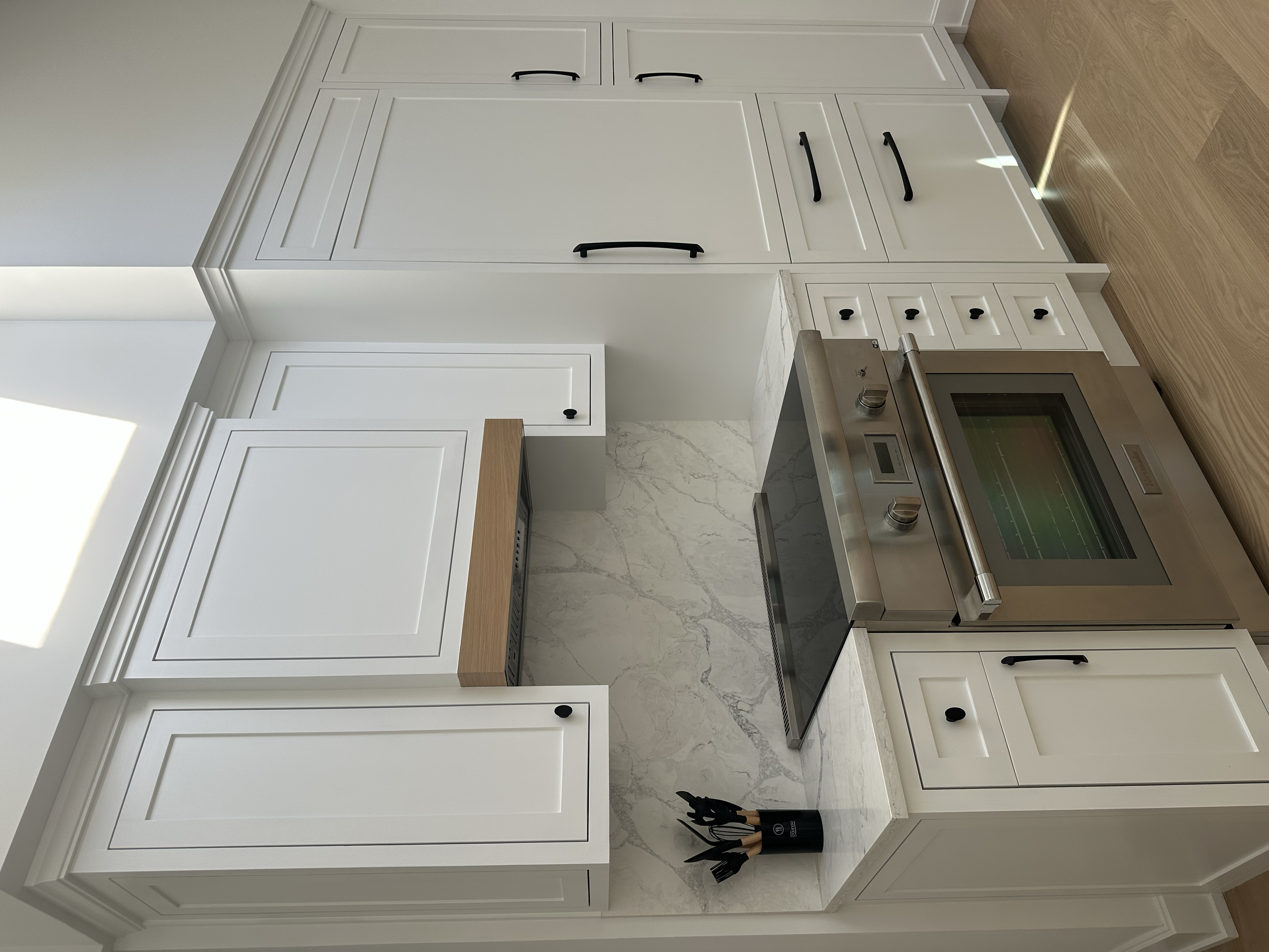 White kitchen cabinets and drawers with black handles, stainless steel oven and stove, marble countertop, and a knife holder with black knives and wooden handles on the countertop.