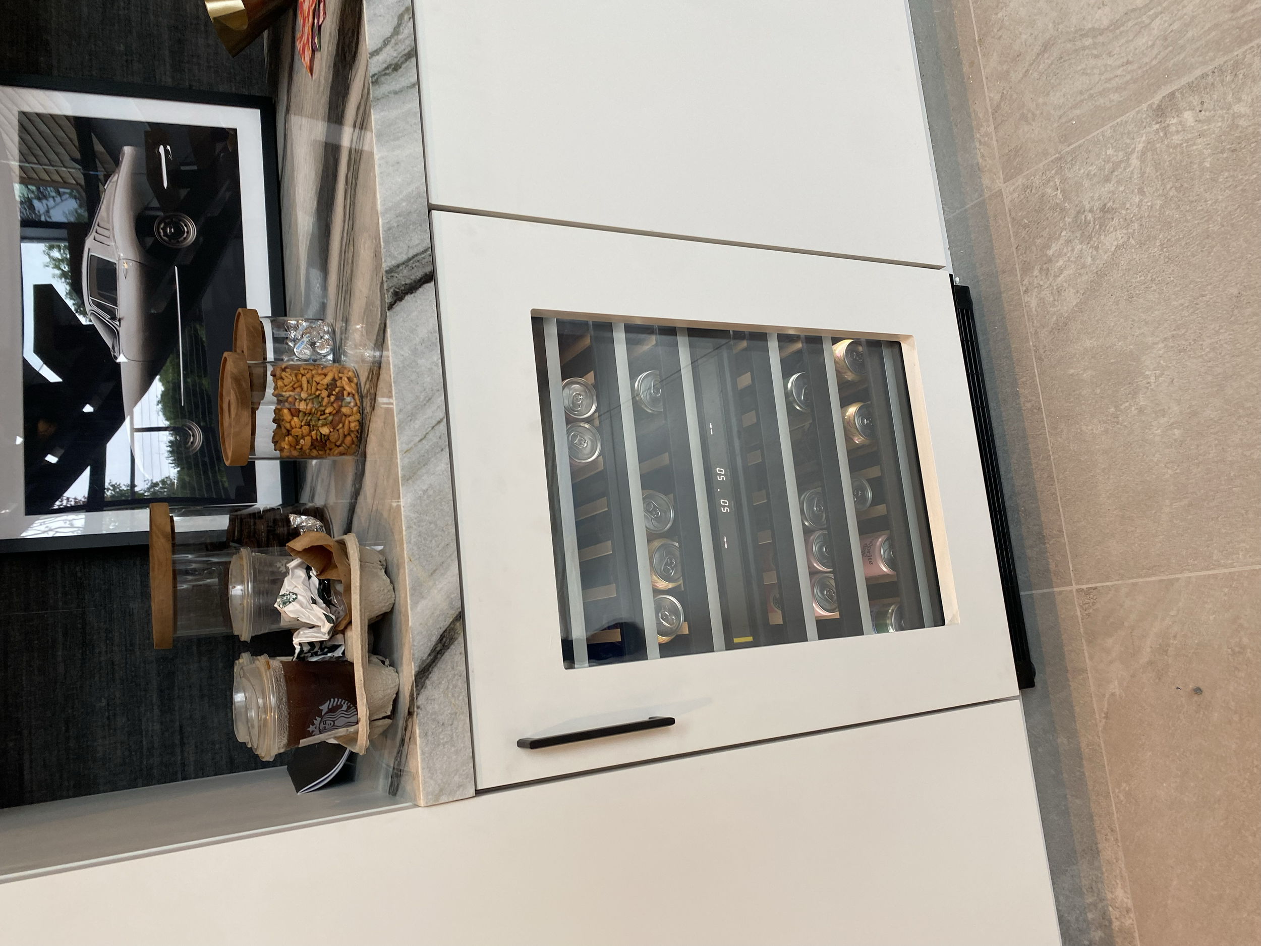 Modern white kitchen cabinet with a built-in wine cooler, countertop with snack jars, glasses, and a framed black-and-white picture of a vintage car.