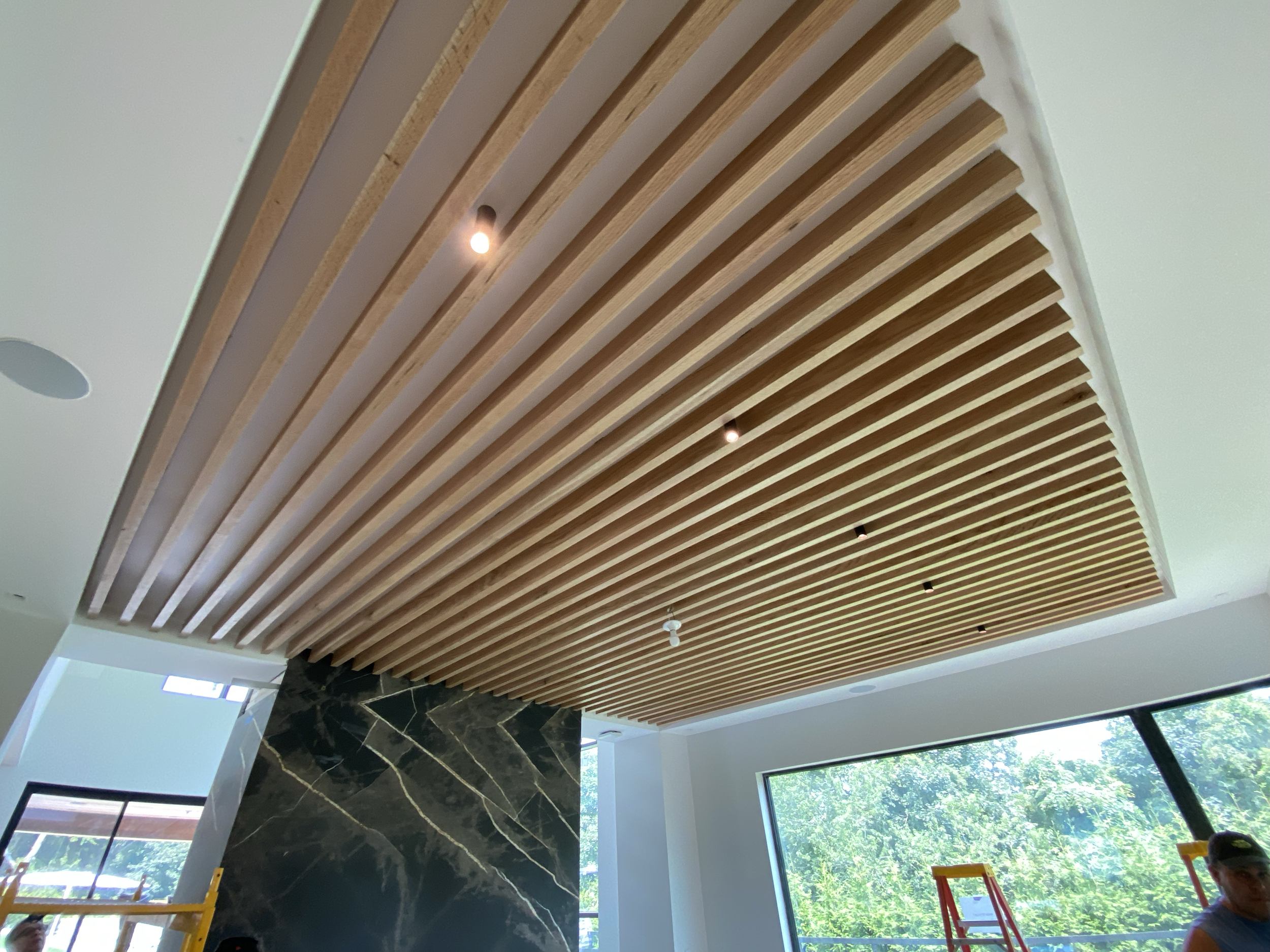 Interior of a building under construction or renovation, showing a ceiling with wooden slats and built-in lighting, large windows letting in natural light, and a marble wall feature.
