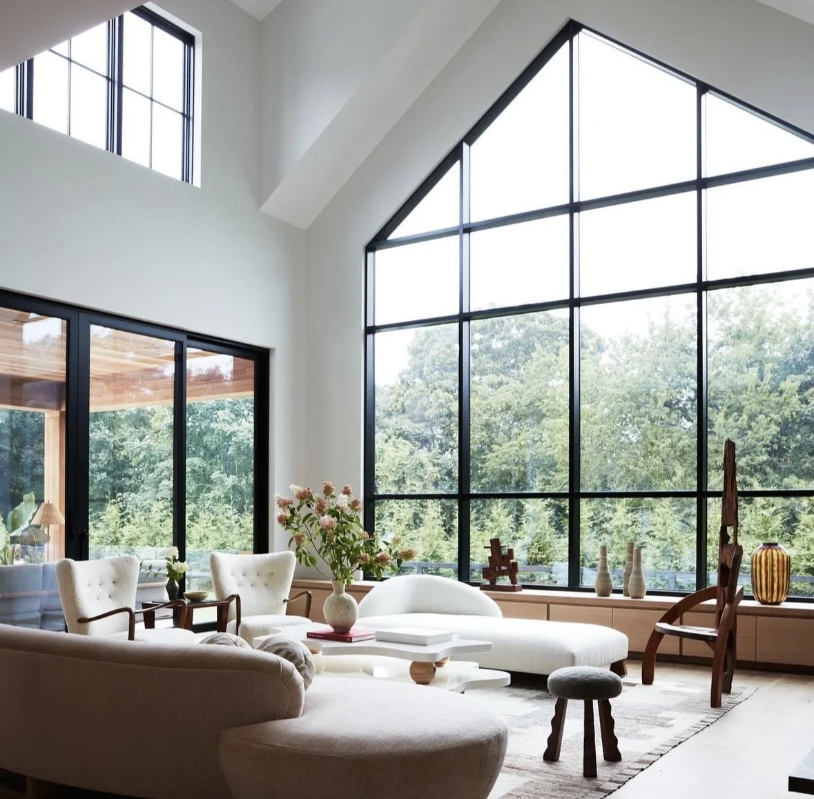 Living room with large floor-to-ceiling windows showing a green outdoor view, beige and white furniture, a vase with pink flowers on the coffee table, and various decorative items on the window ledge. Custom cabinet / storage