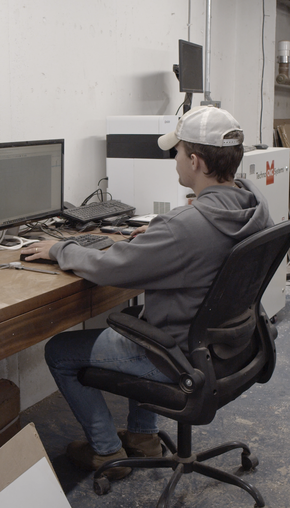 A young man wearing a baseball cap and hoodie seated in a black ergonomic office chair, working at a wooden desk with multiple computer monitors and electronic equipment in a workshop or industrial setting.