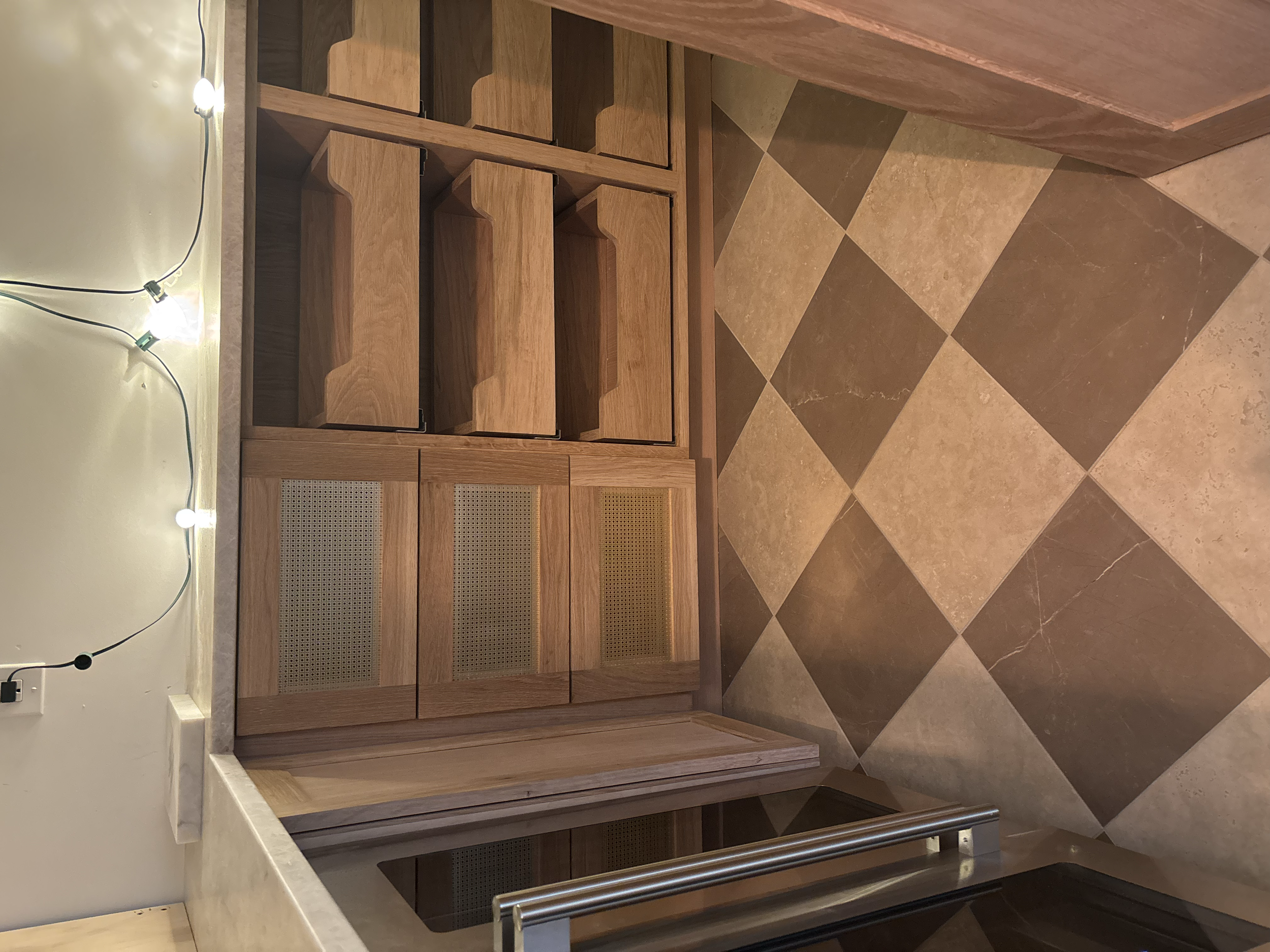 View of a wooden kitchen cabinet with open shelves and rattan doors. The cabinet is next to a wall with a string of decorative lights. The floor is made of large square tiles in beige and dark brown colors.