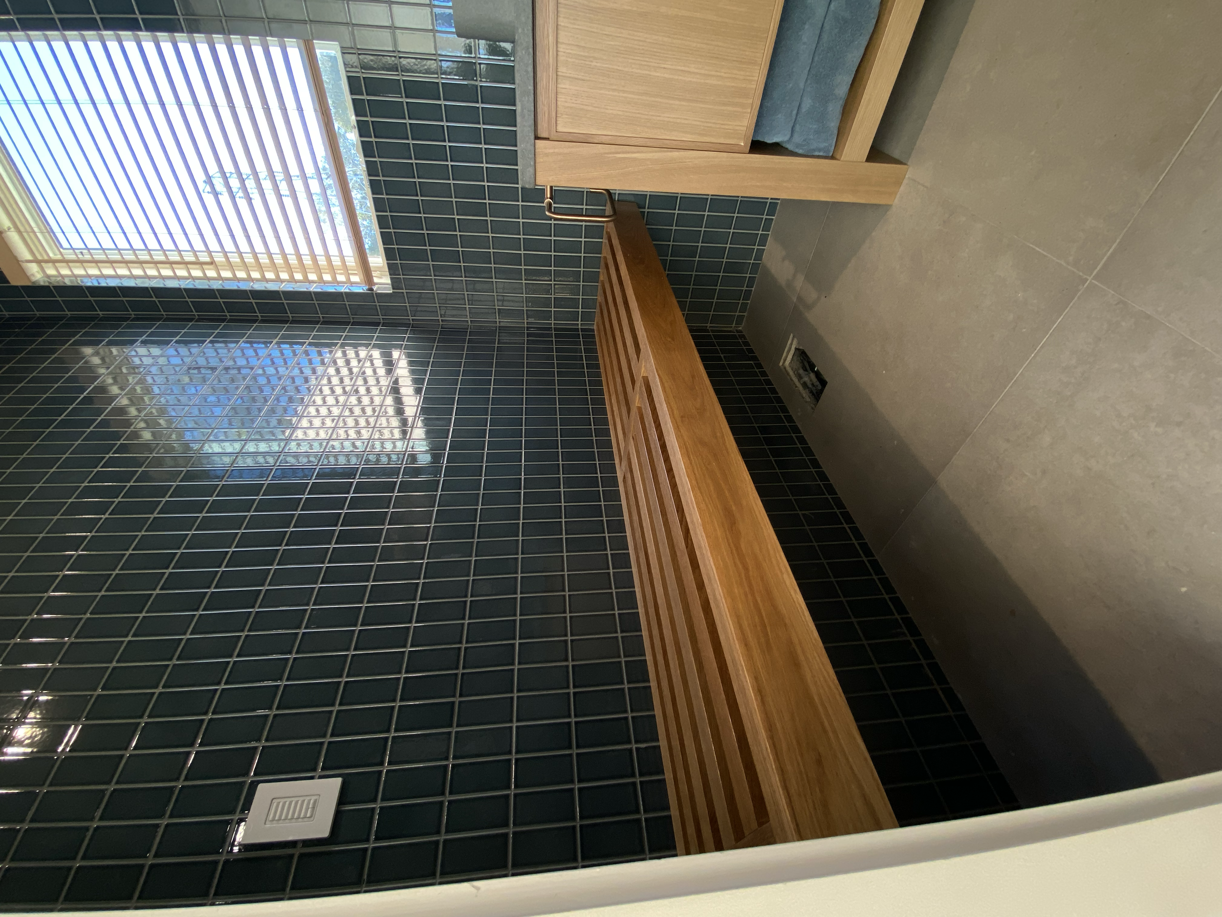 A modern bathroom featuring black tiled walls, a window with blinds, a wooden bench, and a vent.